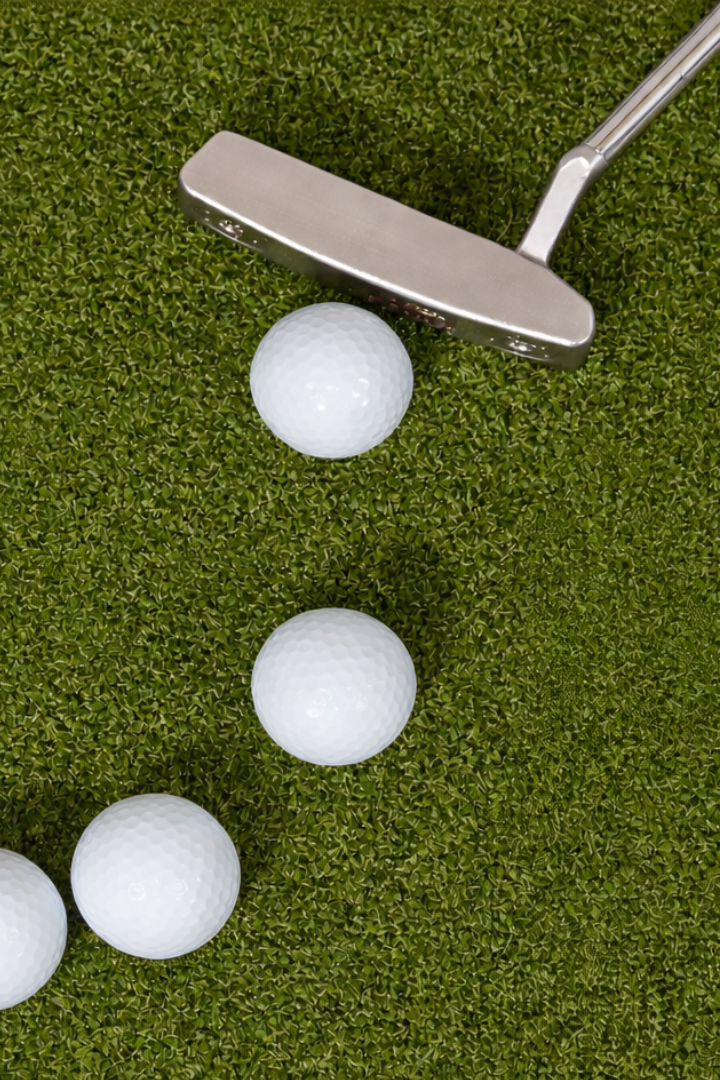 A golf club positioned above three white golf balls on green artificial turf.