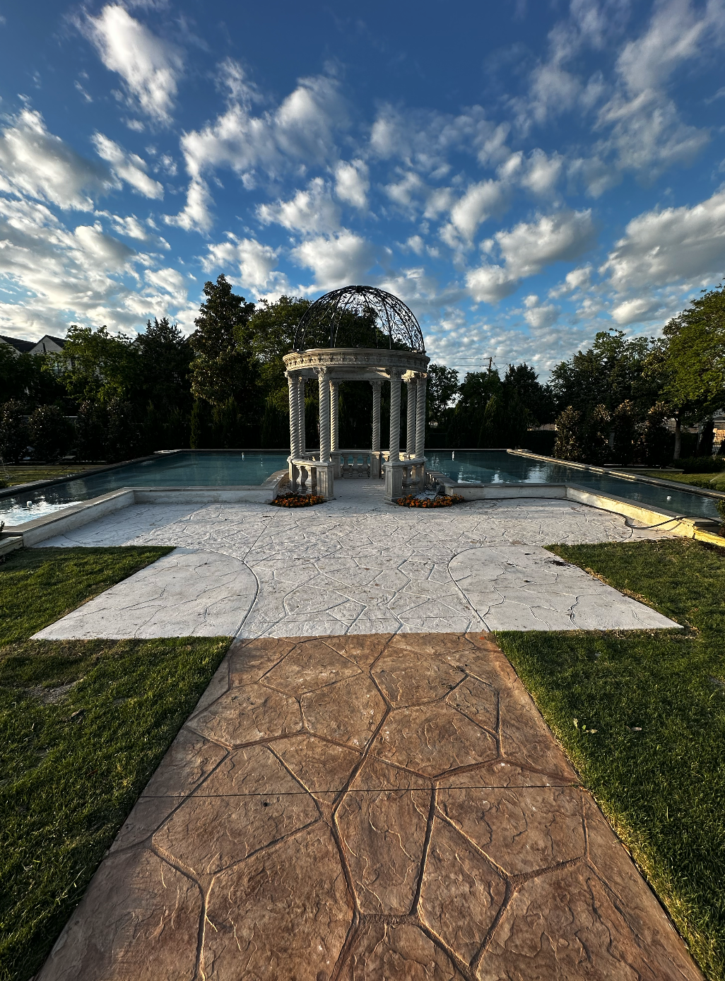 A park scene with a stone pathway leading to a decorative white gazebo at the edge of a swimming pool, under a partly cloudy sky.