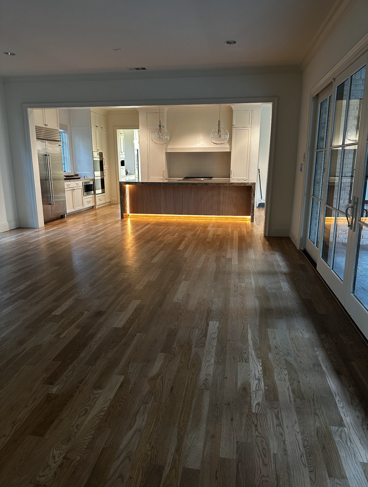 Empty living room with wooden floors, open kitchen in the background, and sliding glass doors on the right.