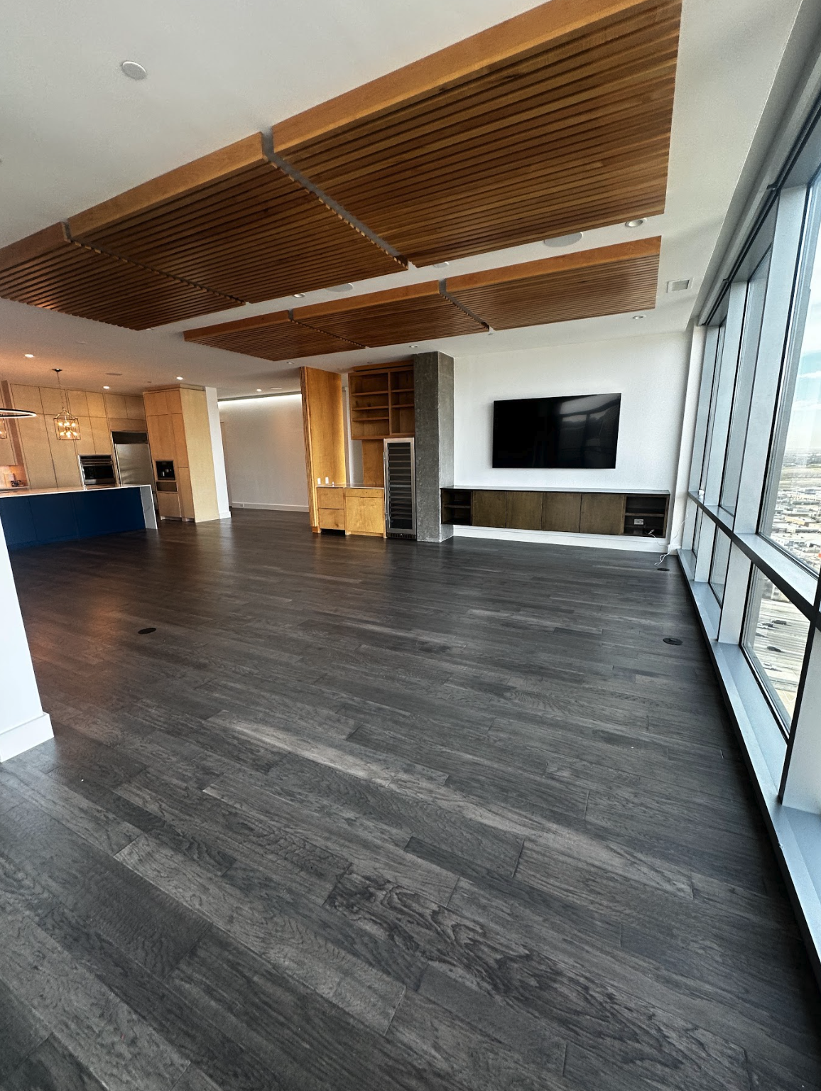 Modern open-concept living space with large windows, dark wood flooring, a wall-mounted TV, wooden ceiling panels, and a kitchen area in the background.