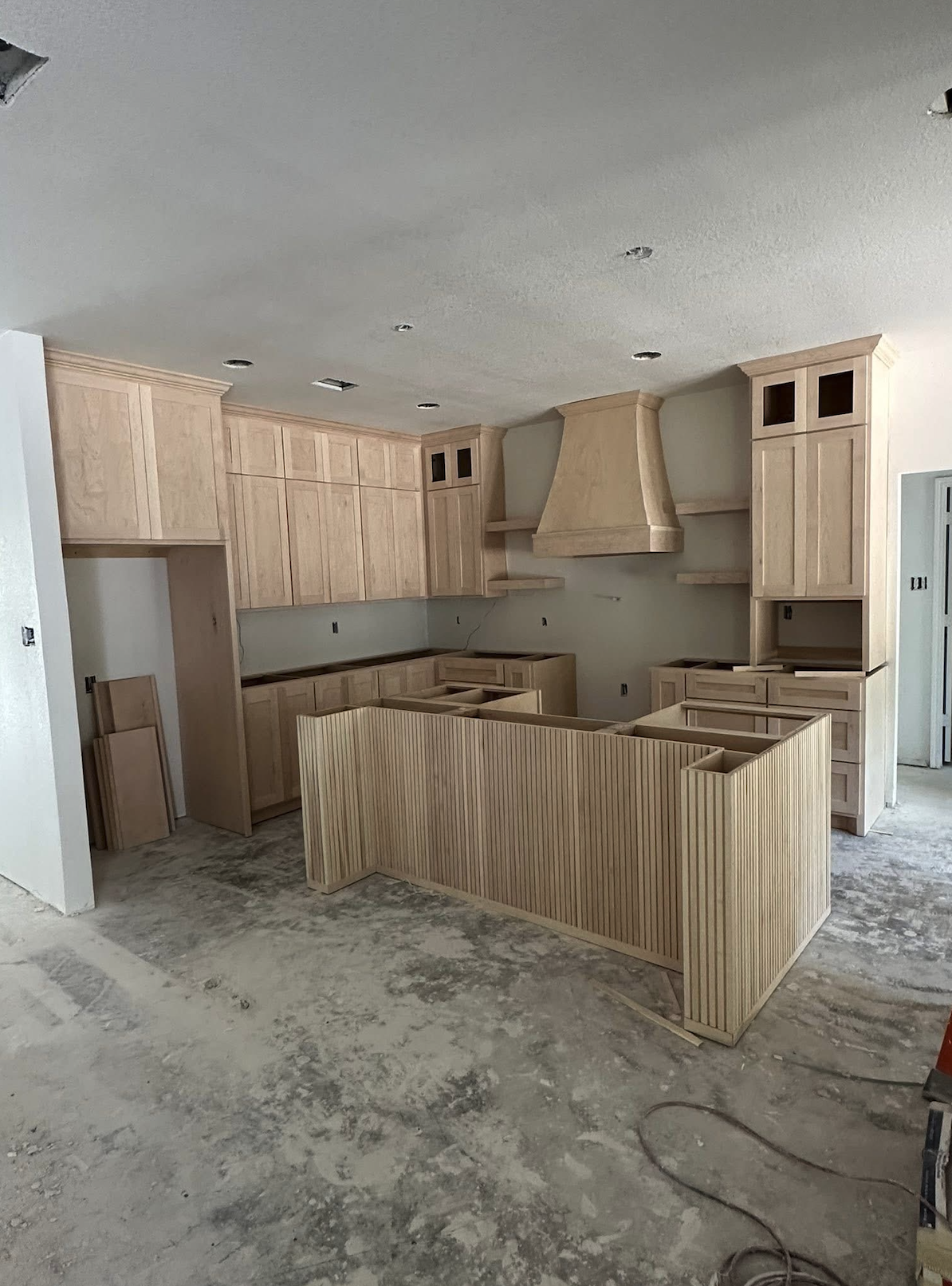 Kitchen under construction with unfinished light wood cabinets, a range hood, and a partially built island, all set on a concrete floor.