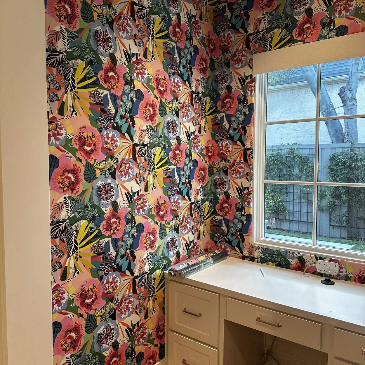 A room with vibrant floral wallpaper, a white countertop, and a window showing a backyard with trees and bushes.