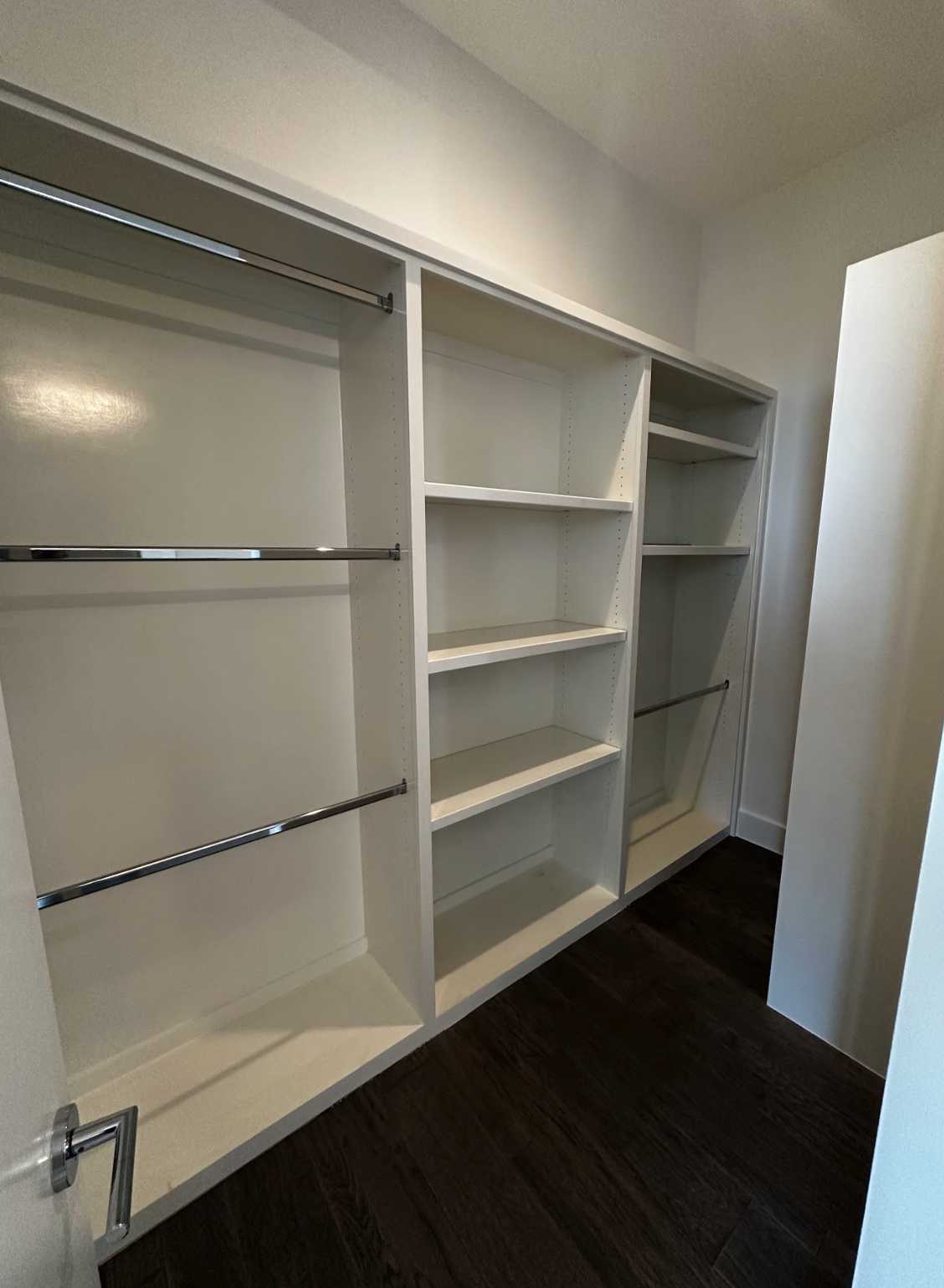 Empty walk-in closet with white shelving and hanging rods, dark hardwood floor, and beige walls.