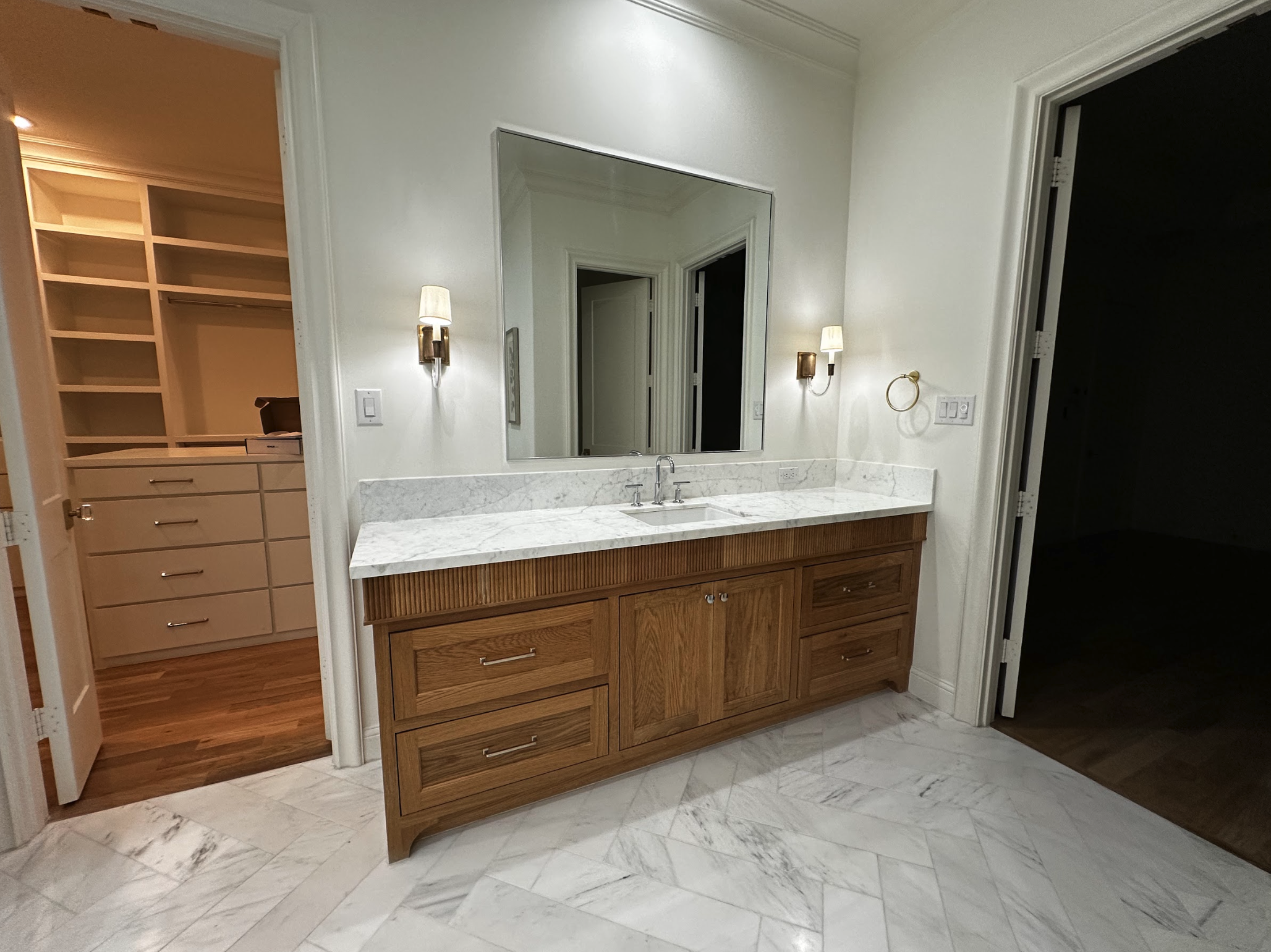 Bathroom vanity with a marble countertop, wooden drawers, and a large mirror. On the wall, there are two wall-mounted lights, a towel ring, and a switch. To the left, an open closet with wood shelves and drawers is visible. The floor has marble tiles
