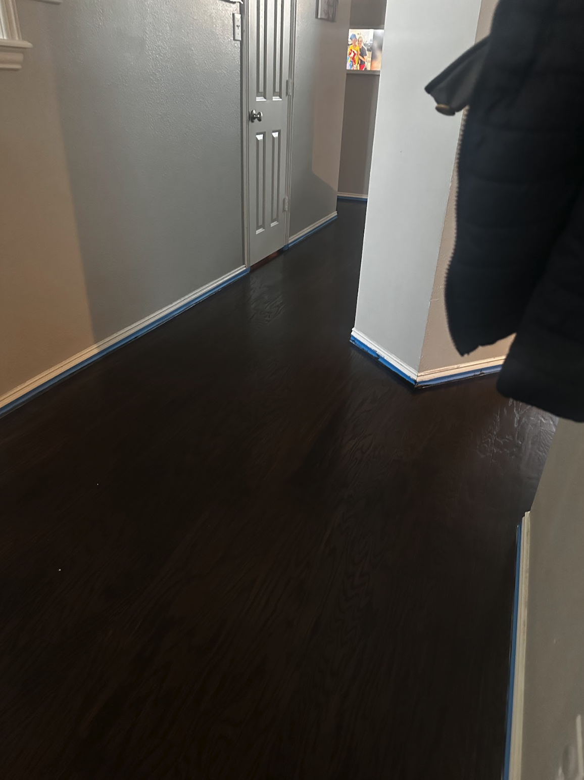 Interior hallway with dark hardwood floors, beige walls, a closed door, and blue painter's tape along the baseboards and edges of the wall.