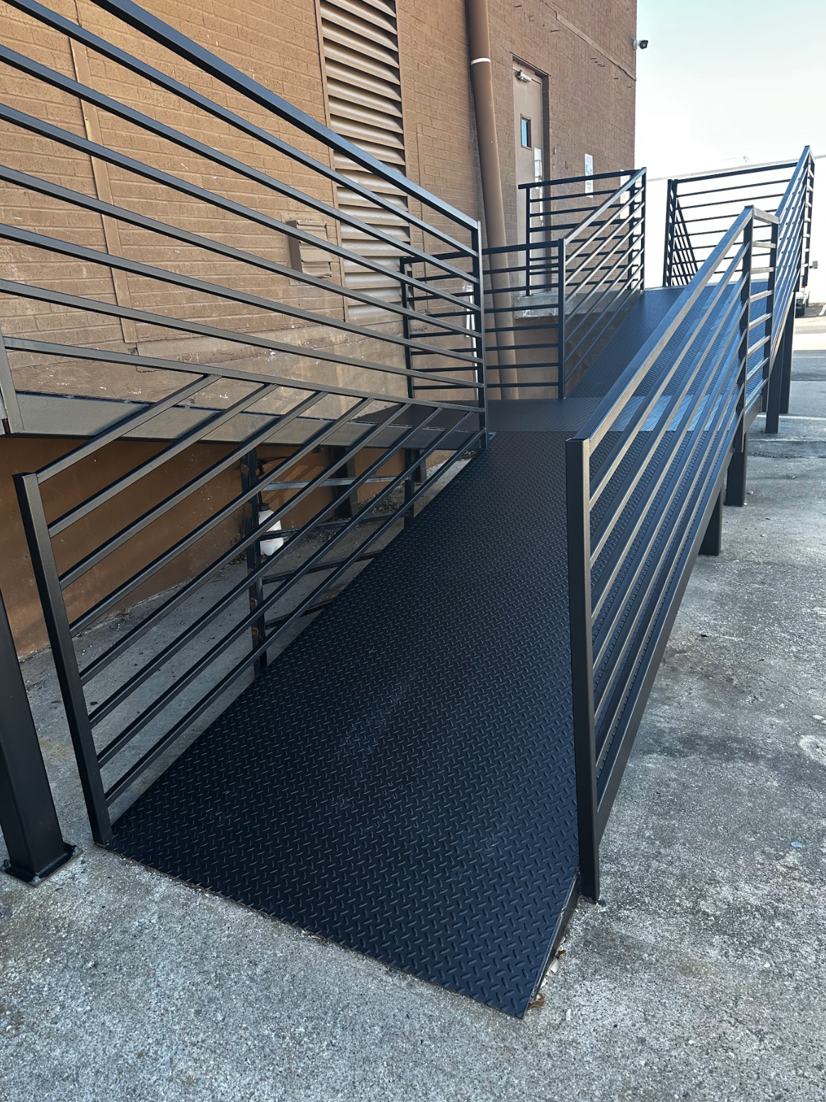A black metal wheelchair ramp with railings on both sides, leading up to a building entrance. The building has a brown brick exterior with ventilation louver and a brown drainpipe. The ramp is made of diamond plate metal for slip resistance.