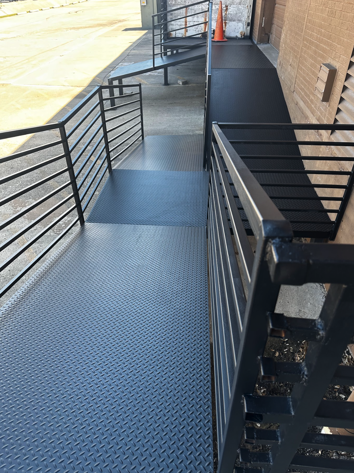Metal ramp with black railings outside a brick building, with an orange safety cone at the top, leading to a door.