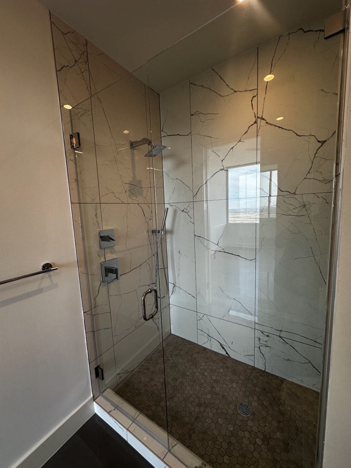 A modern shower with marble walls, glass door, and brown hexagonal tile flooring, featuring a city view through a nearby window.
