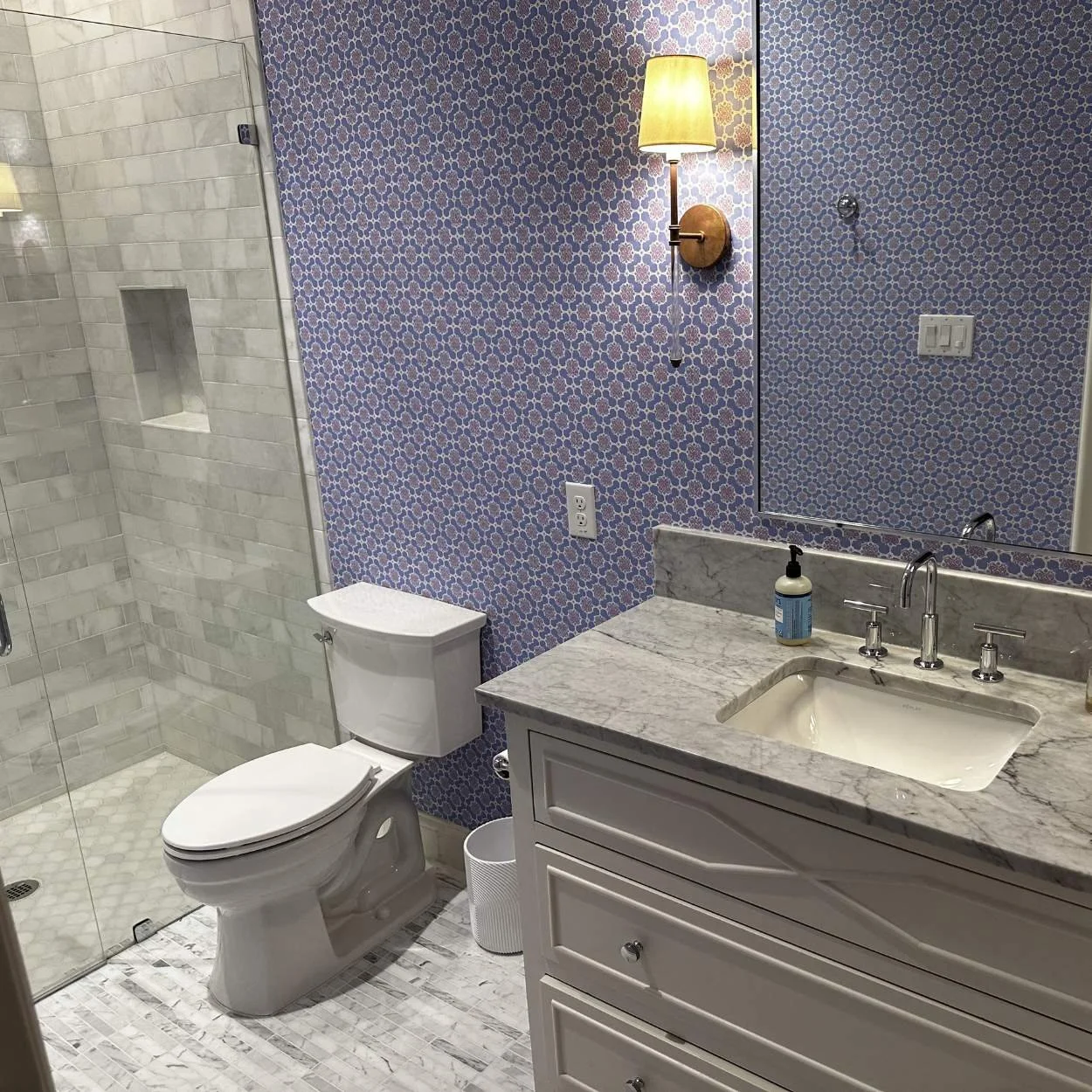 A bathroom with patterned wallpaper, a wall-mounted light, a mirror, a marble countertop with a sink, a soap dispenser, a toilet, and a glass-enclosed shower with white subway tiles and a built-in niche.