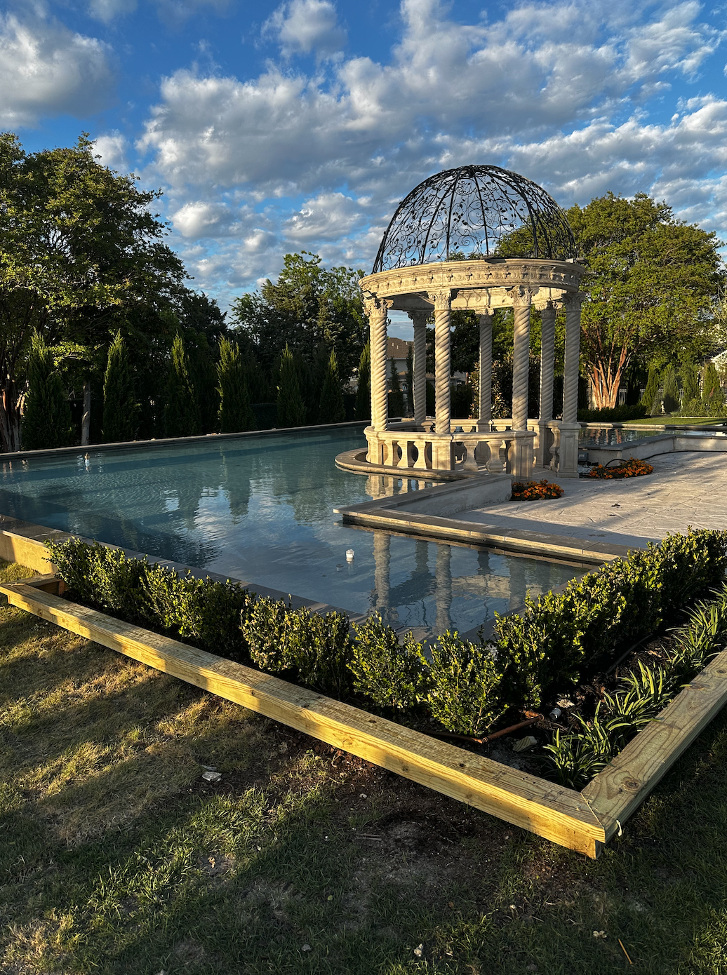 A decorative pool with a classical-style gazebo featuring twisted columns and a wrought iron dome, surrounded by greenery and flowers, under a partly cloudy sky.