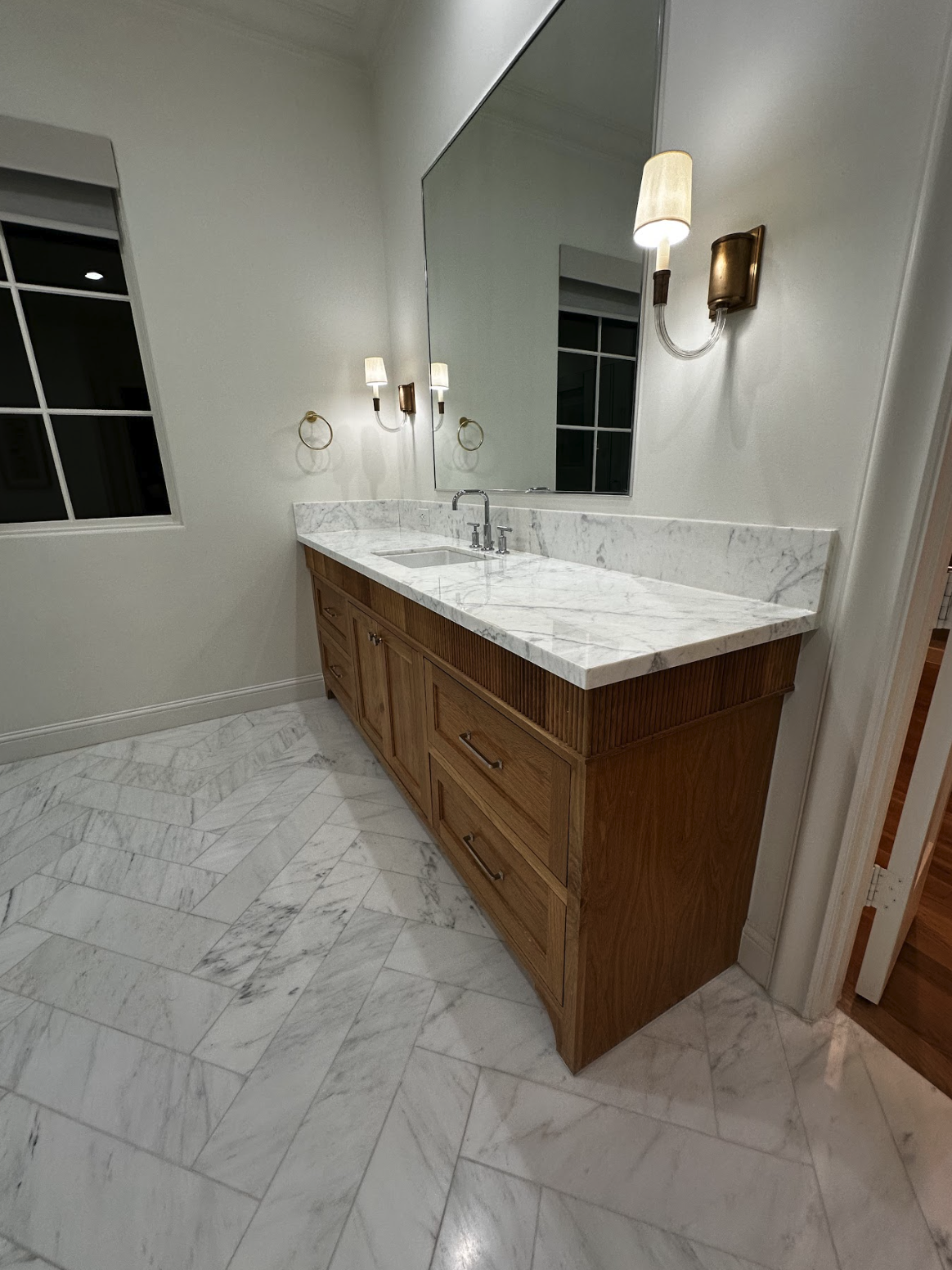 Bathroom with a wooden vanity, white marble countertop, large mirror, wall-mounted lamps, and a window with a white frame.