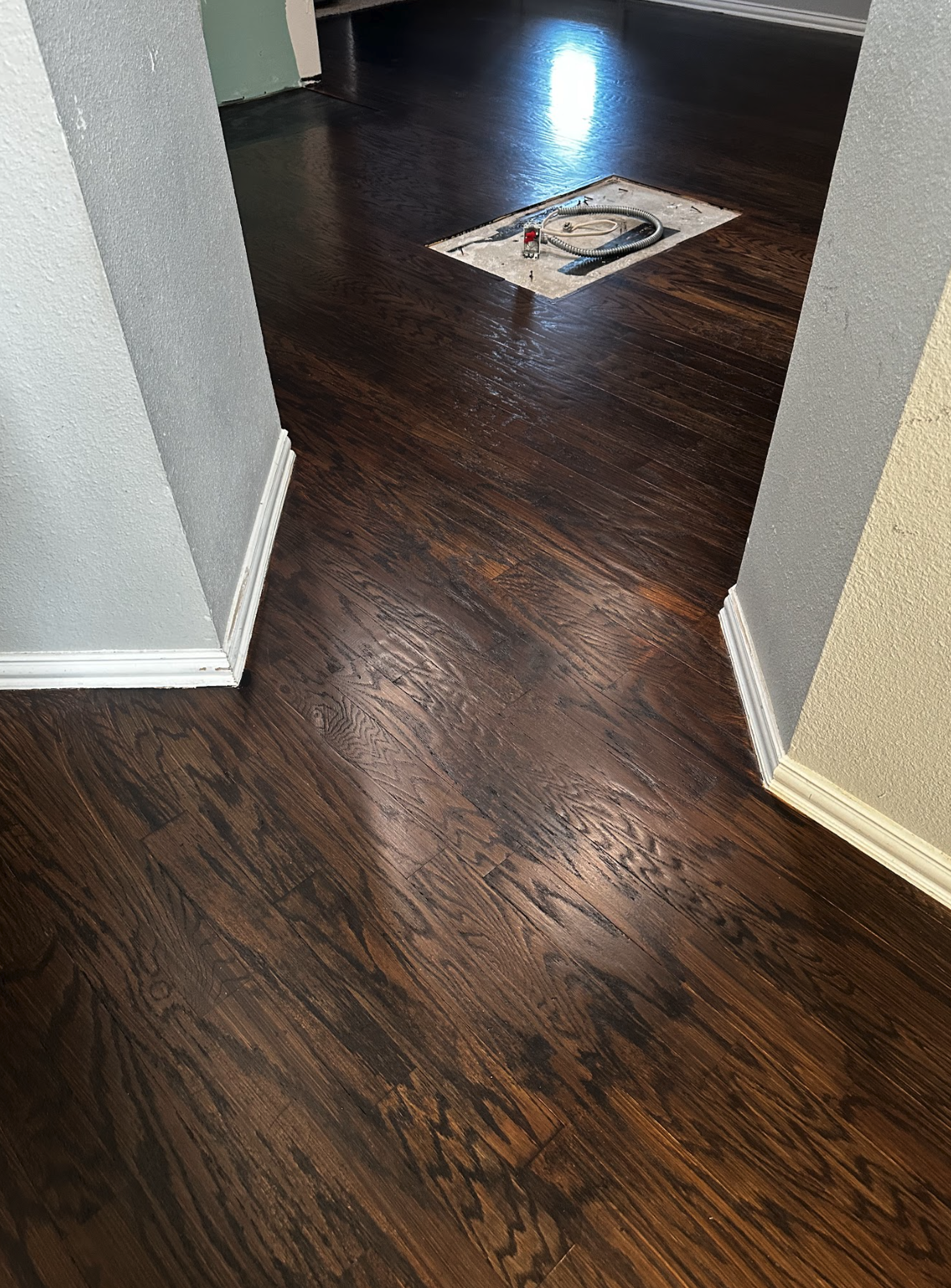 Dark hardwood flooring in a room with light-colored walls, an open door, and a covered hole in the floor with a vacuum hose and electrical wires.