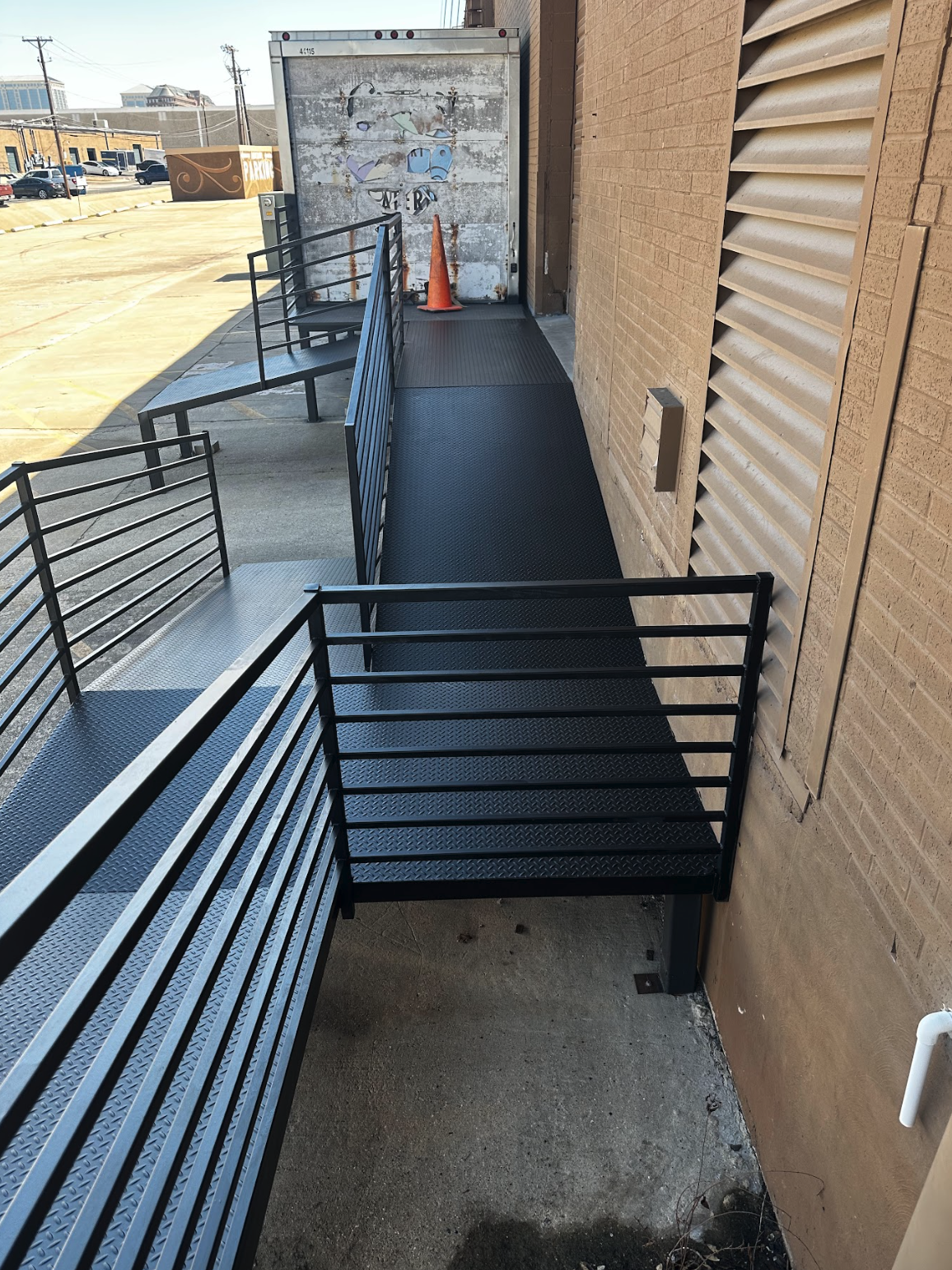 Exterior metal ramp with black railings next to a brick building, leading to a back door with a vent and a parking lot in the background.