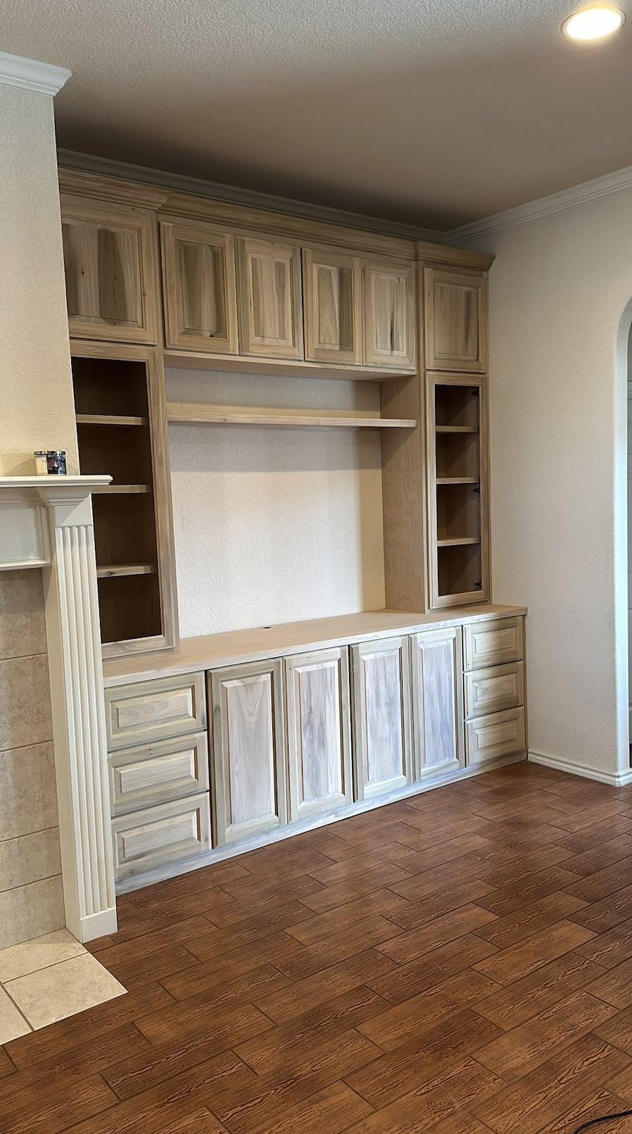 Empty built-in wooden shelving and cabinet unit in a room with hardwood flooring and a partial view of a fireplace.