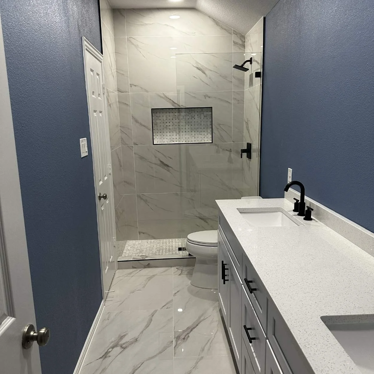 Modern bathroom with white marble flooring, blue walls, a white vanity with black handles, a black faucet, and a glass-enclosed shower with marble tiles and a small built-in shelf.