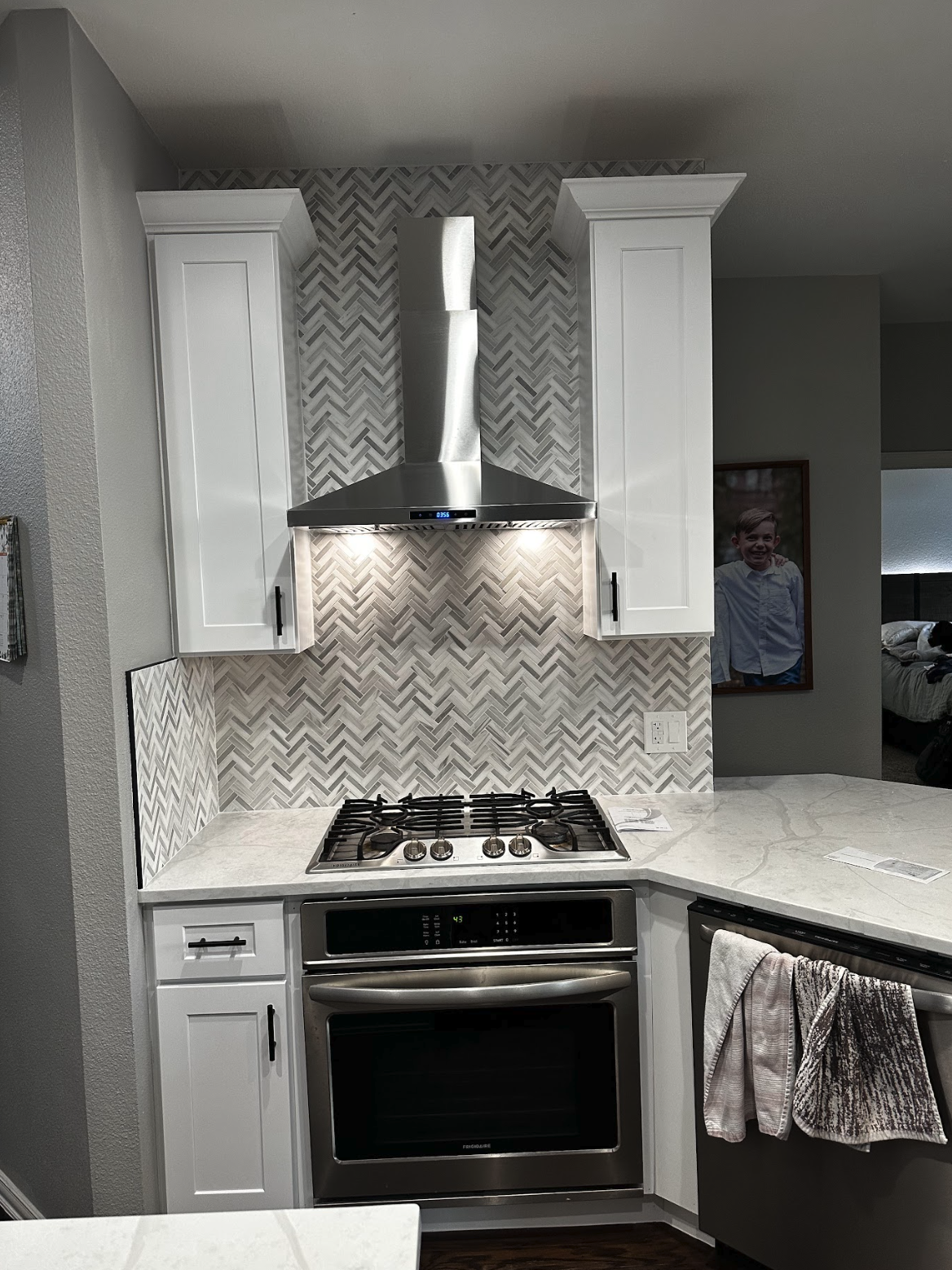 Modern kitchen with white cabinets, a stainless steel range hood, a marble countertop, and a stainless steel oven. There is a patterned tile backsplash behind the stove and some towels hanging on the oven handle. A framed photo of a child is visible 