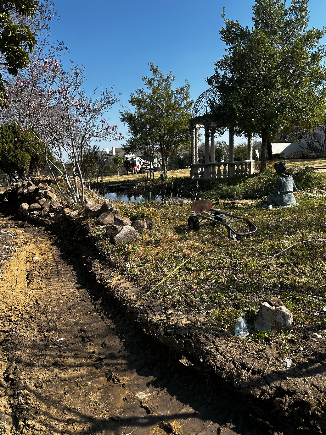 A garden scene with a small pond, trees, a white stone gazebo, and a statue of a girl sitting on the grass, with a lawn mower lying on the grass and a dirt path in the foreground.