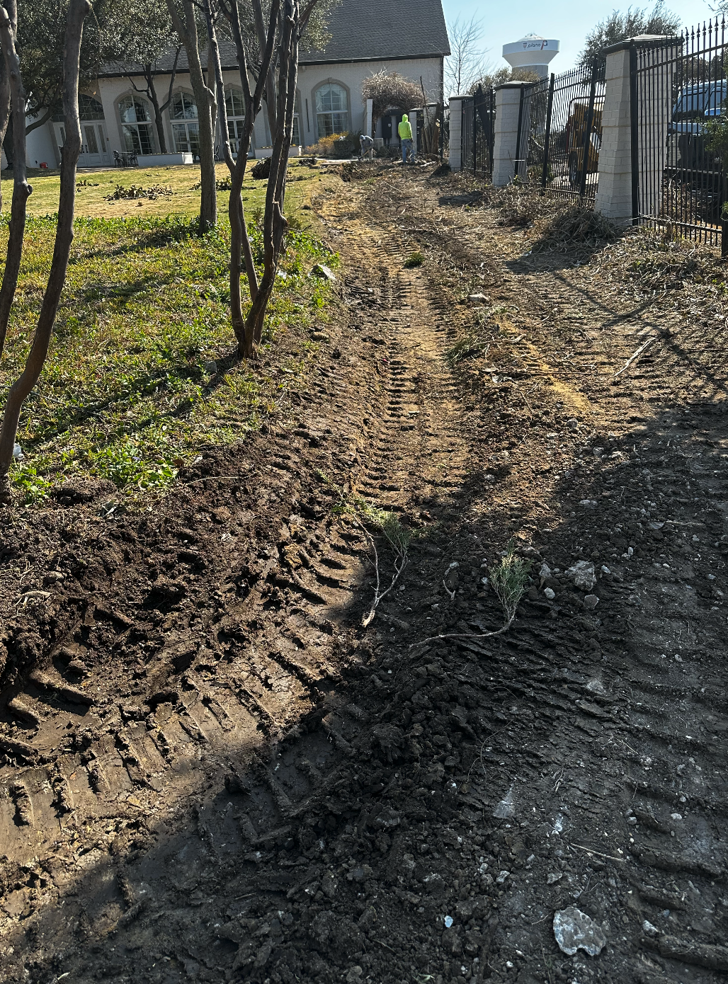 Dirt pathway with tire tracks near a fenced yard and a house, with workers in the background landscaping.