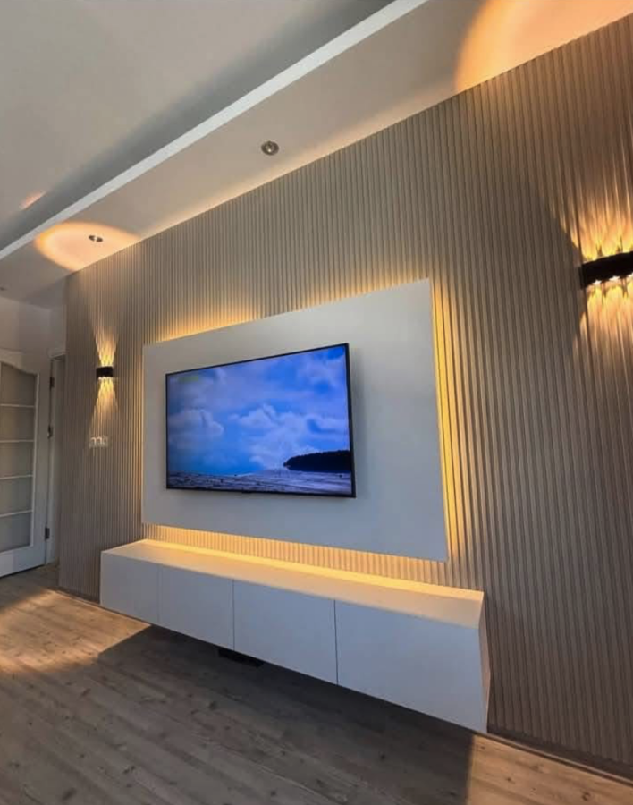 A modern living room with a flat-screen TV mounted on a wall with wooden paneling. The TV displays a landscape with clouds and a body of water. There is a white, minimalist floating cabinet below the TV, and warm lighting accents the wall. The floor 