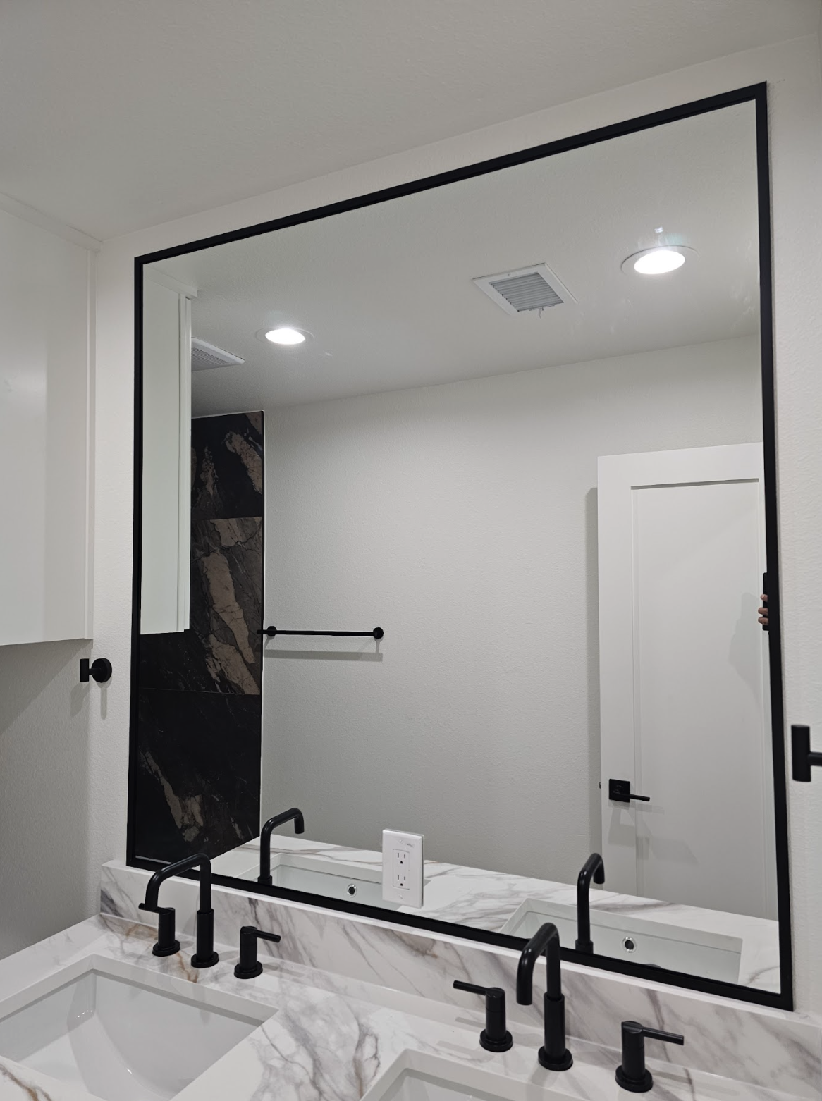 A large bathroom mirror reflecting a white wall with a towel bar, a black marble accent, and ceiling lights, above a marble countertop with sinks and black faucets.
