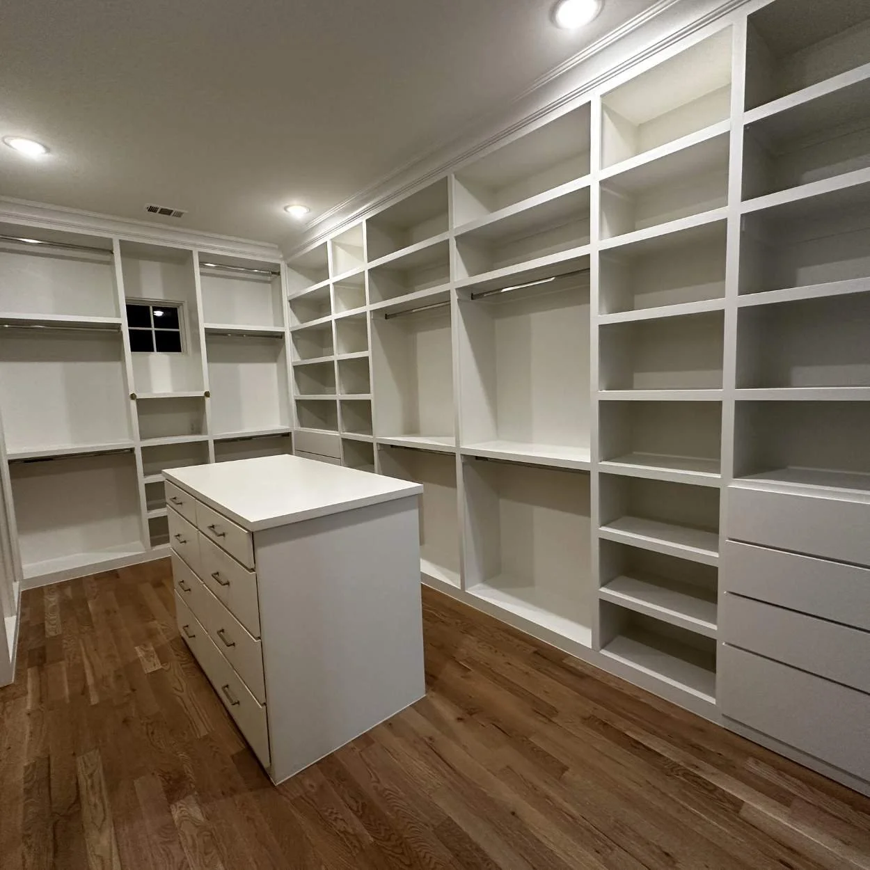 Empty walk-in closet with white built-in shelves, drawers, and an island island in the center, with hardwood flooring and recessed lighting.