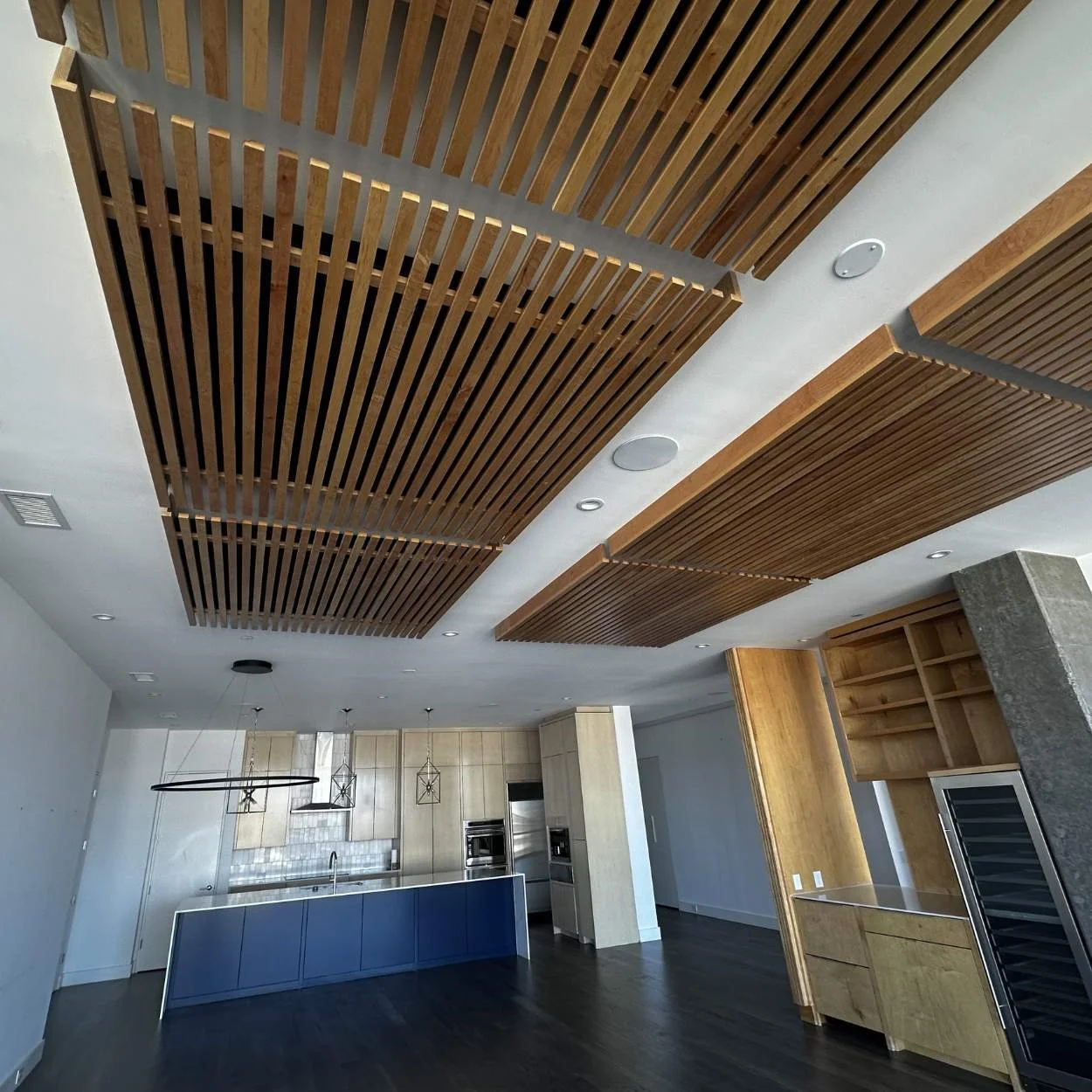 Modern kitchen with wooden slatted ceiling panels, light-colored cabinetry, and a dark hardwood floor.