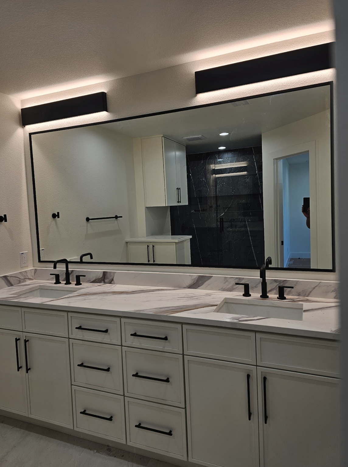 A modern bathroom with a large mirror above a white vanity with black handles and fixtures, a marble countertop, and a view of a separate shower area with black marble walls.