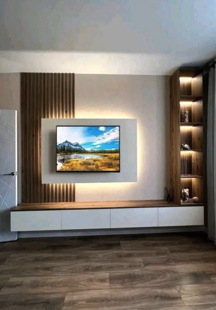 Modern living room with mounted flat-screen TV, wooden panel behind it, white entertainment unit below, built-in wooden shelving with decorative items to the right, and light-colored walls and flooring.