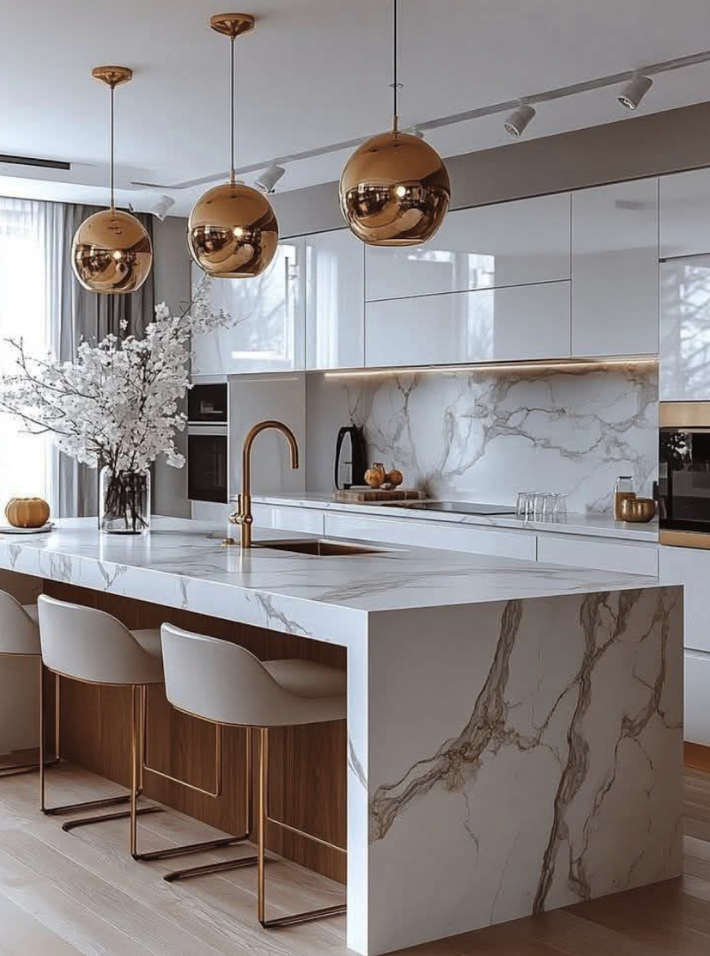 Modern kitchen with white marble countertops, a marble backsplash, and gold accents. Three gold pendant lights hang above the island, which has a vase with white flowers and a pumpkin. There are four beige barstools with gold legs.