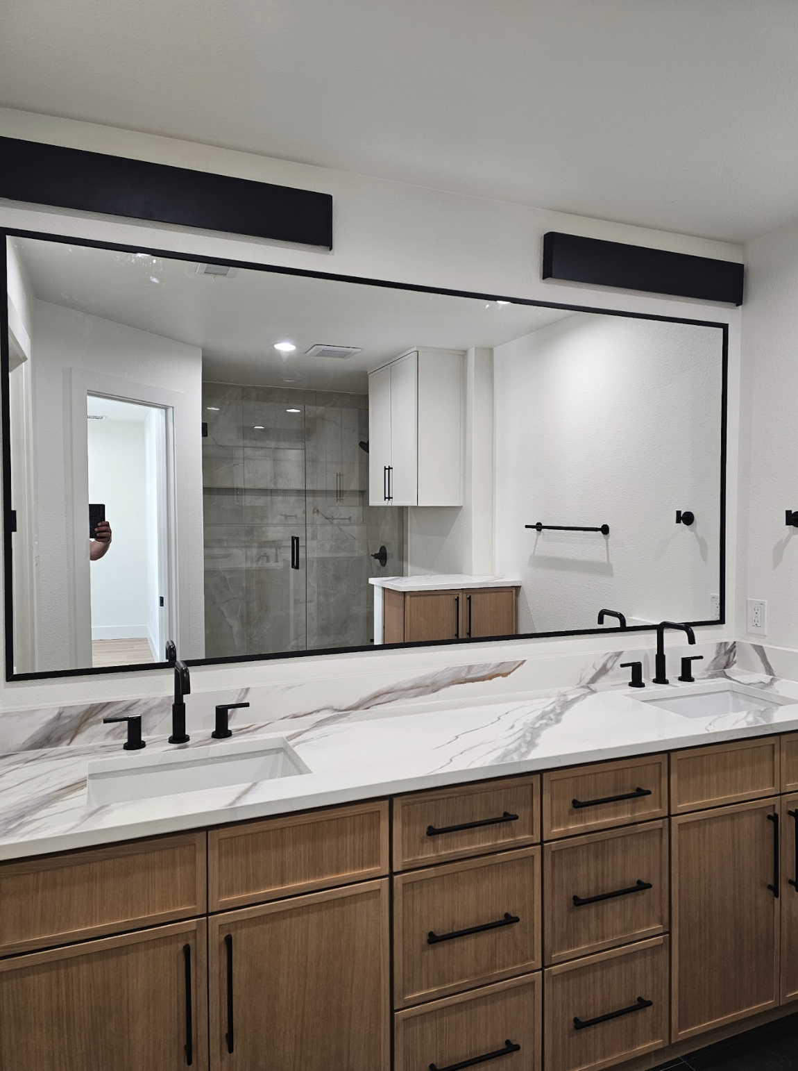 Bathroom vanity with a large mirror, white marble countertop with brown veining, black faucets and hardware, wooden cabinets, visible bathroom shower with glass door and tiled walls, and a black towel bar.