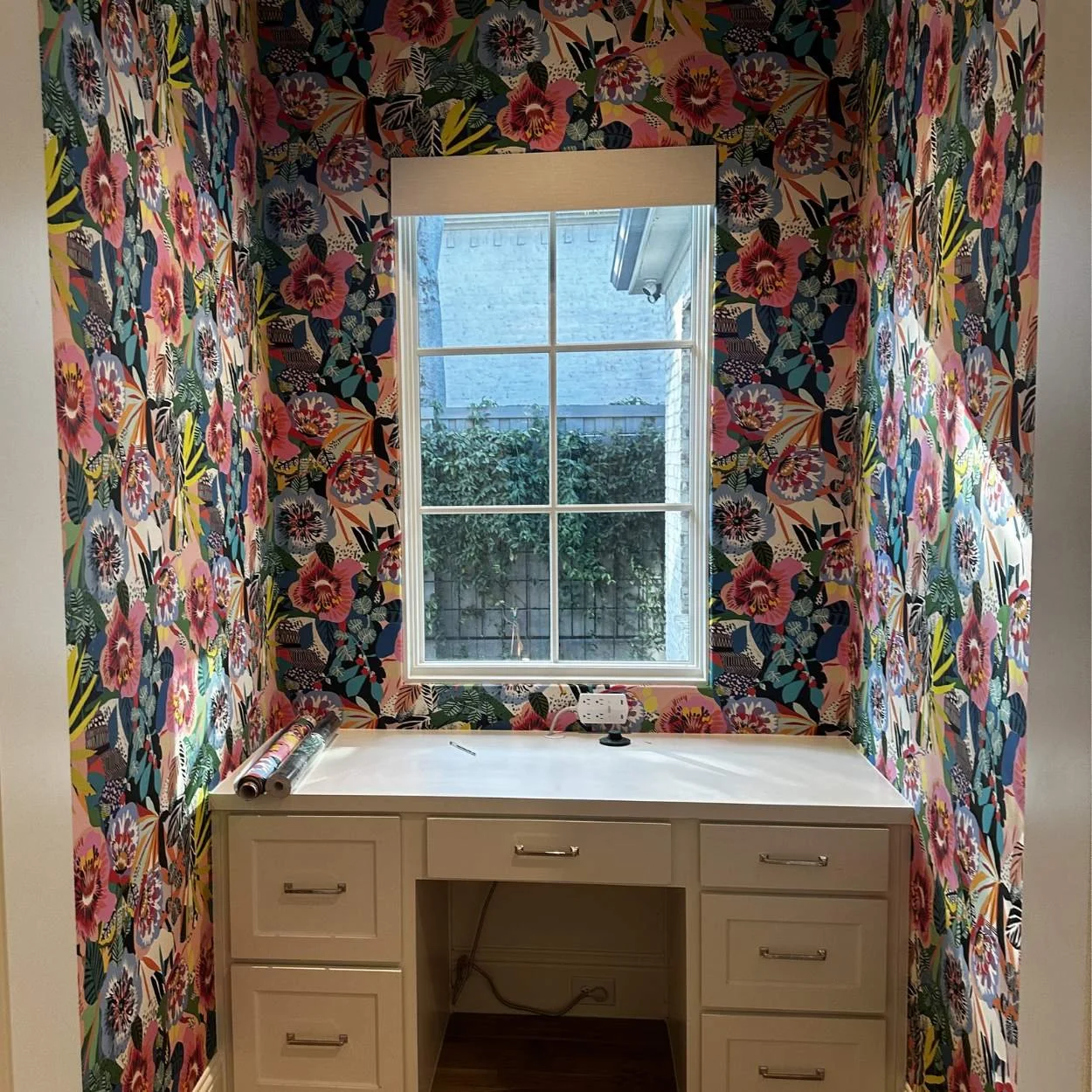 A bright room with floral wallpaper on all walls, featuring pink hibiscus and green leaves. A white desk with drawers is positioned under a window with a beige valance. On the desk, there are a pair of rolled-up fabrics, an electrical outlet, a black