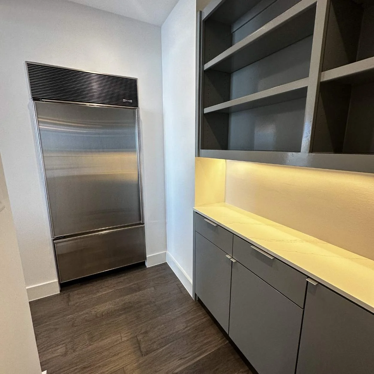 Modern kitchen with stainless steel fridge, gray cabinetry, open shelves, and a yellow-lit counter