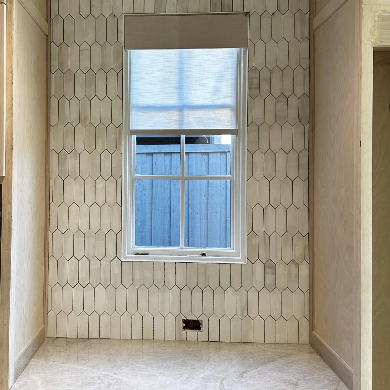 Interior wall with hexagonal tile pattern, a window with a partially closed shade, and plumbing opening at the bottom.