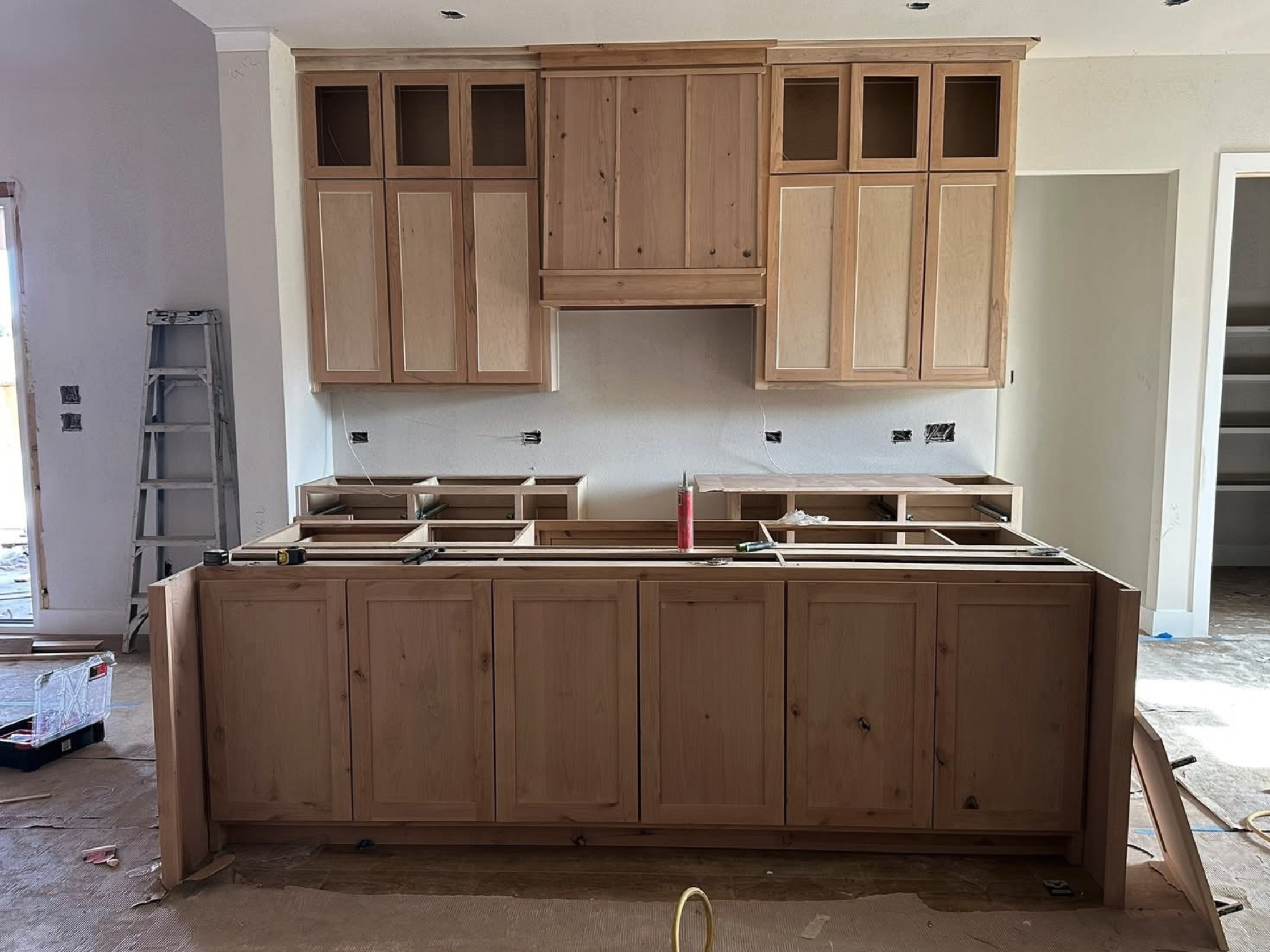 Wooden kitchen cabinets under construction in a room with unfinished walls and exposed outlets.