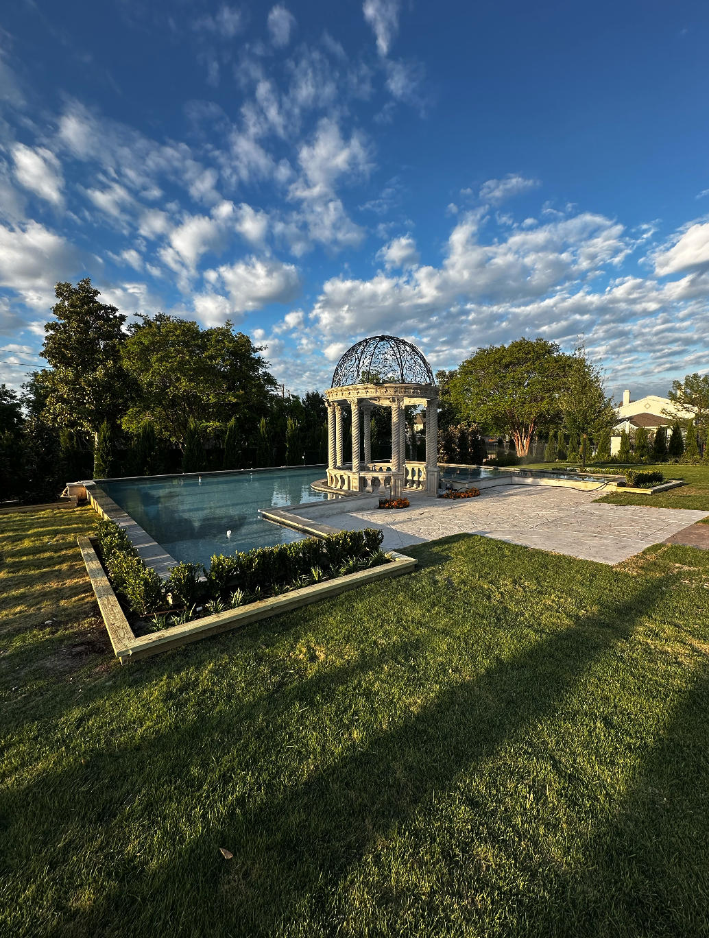 A decorative pool with a white gazebo structure with a metal dome on top, surrounded by grass, trees, and a partly cloudy blue sky.