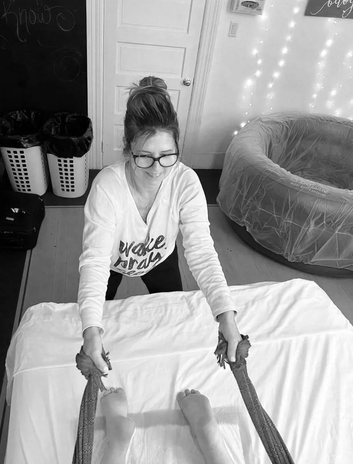 A woman with glasses and a messy bun is smiling as she holds the feet and ankles of someone lying on a bed, with the person's legs extended towards her. The room has fairy lights on the wall, a large round inflatable chair, and laundry baskets in the