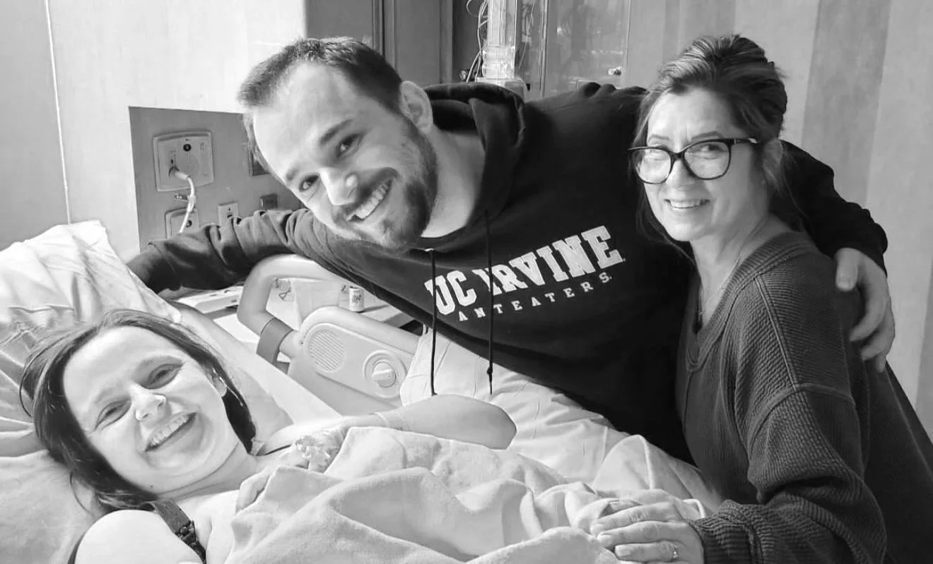 A woman in a hospital bed smiling with a bandage on her arm. Two people, a man and a woman, stand next to her, smiling and looking at the camera. The man is wearing a hoodie, and the woman has glasses and short hair.