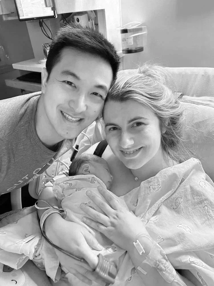 A new mother in a hospital bed holding a newborn baby, with a smiling father leaning in next to her.