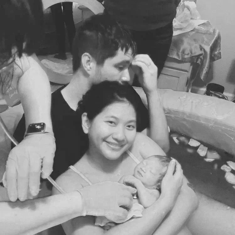 A woman holding a newborn baby in a hospital room, surrounded by medical staff. The woman is smiling, and the baby is getting cleaned or checked by the medical staff.