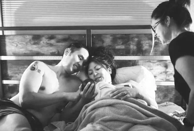 A man and woman in bed looking at a phone together, with another woman sitting nearby, all smiling.