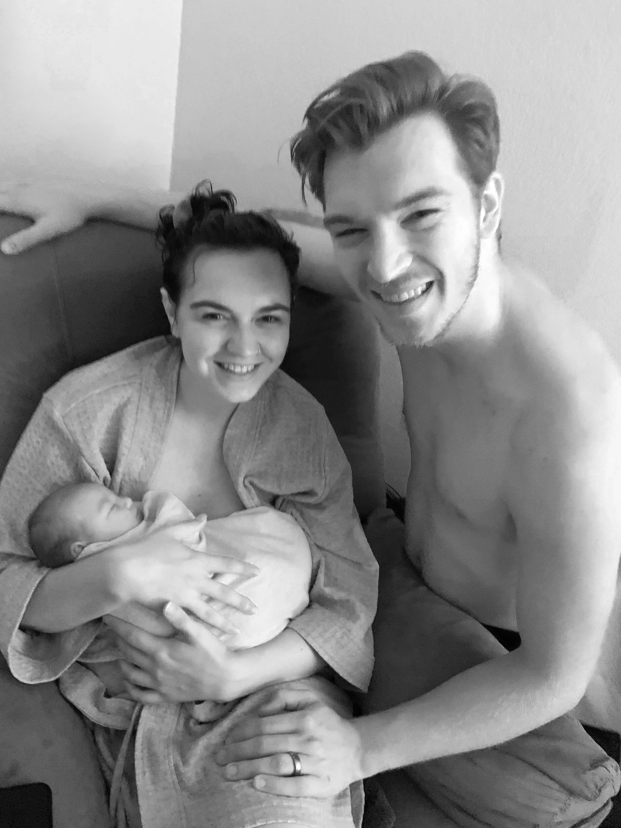 A couple with a newborn baby, the woman is sitting and holding the baby, smiling, while the man stands next to them, shirtless, smiling, with one hand on the woman's knee.