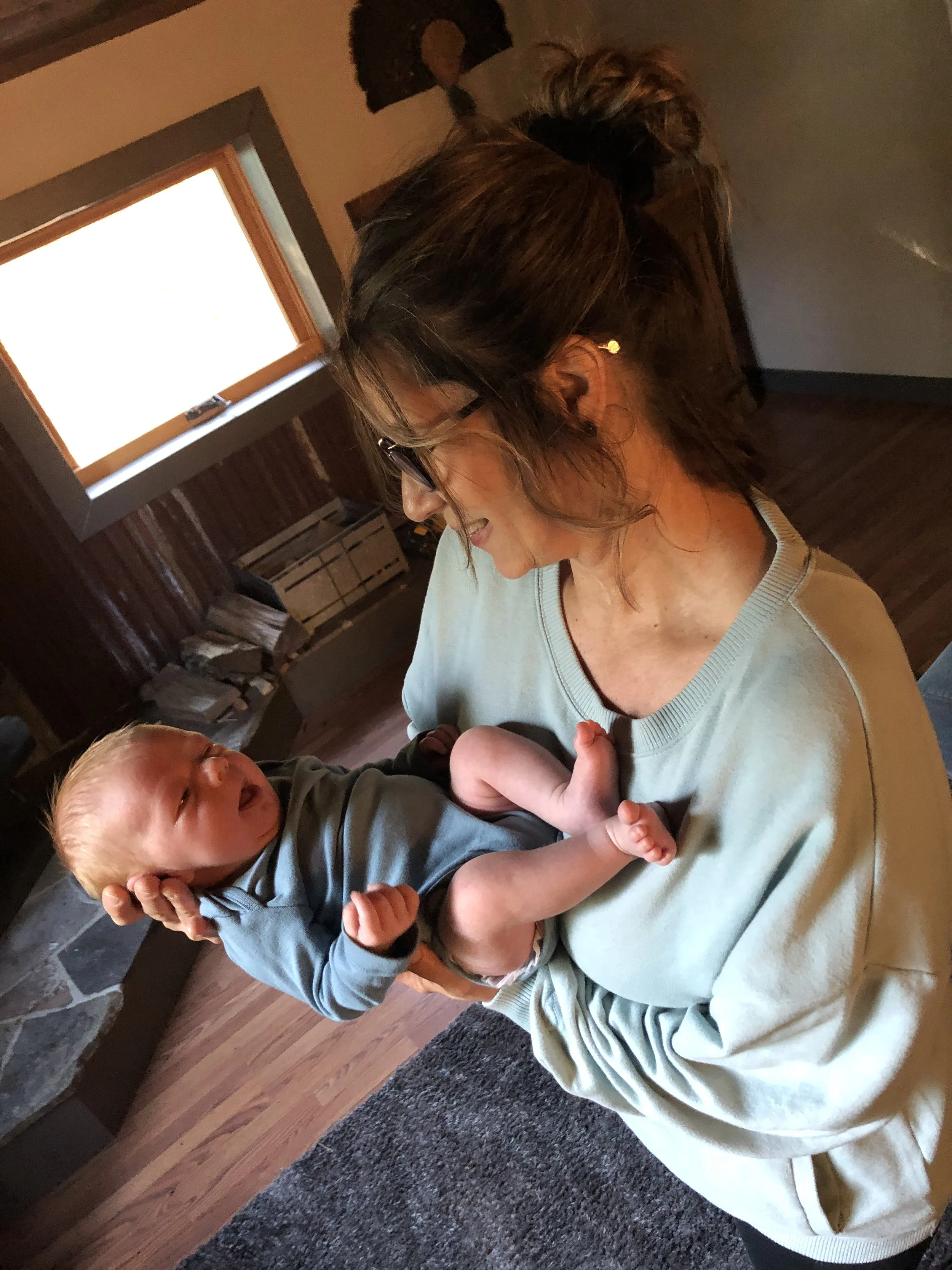 Yoli with dark hair in a messy bun and glasses is smiling and holding a crying baby with light hair, dressed in a long sleeve shirt.