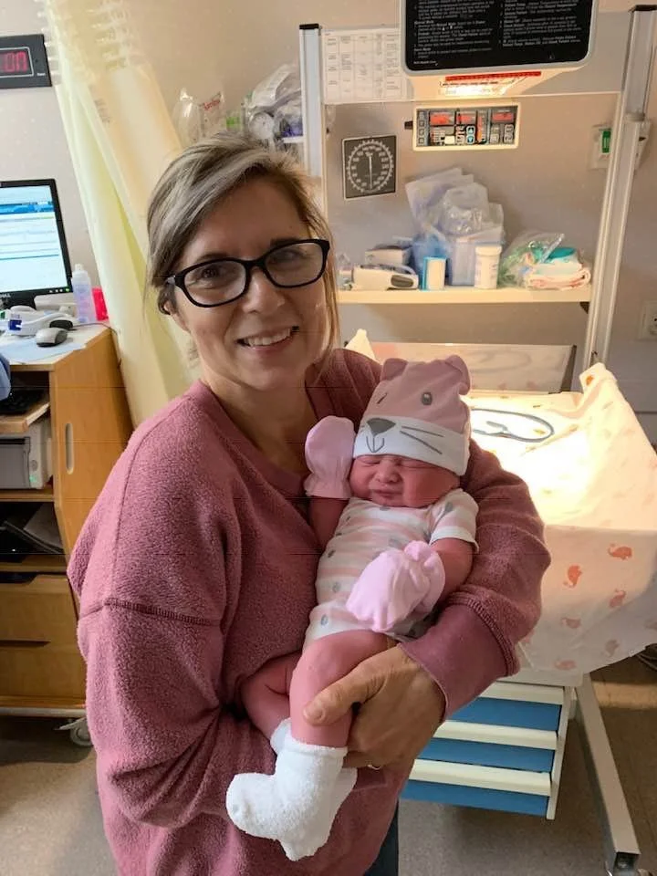 Yoli holding a newborn baby in a hospital room. The woman is smiling and wearing glasses and a pink sweater. The baby is dressed in a striped onesie, wearing a pink hat with a cat face design and pink mittens.