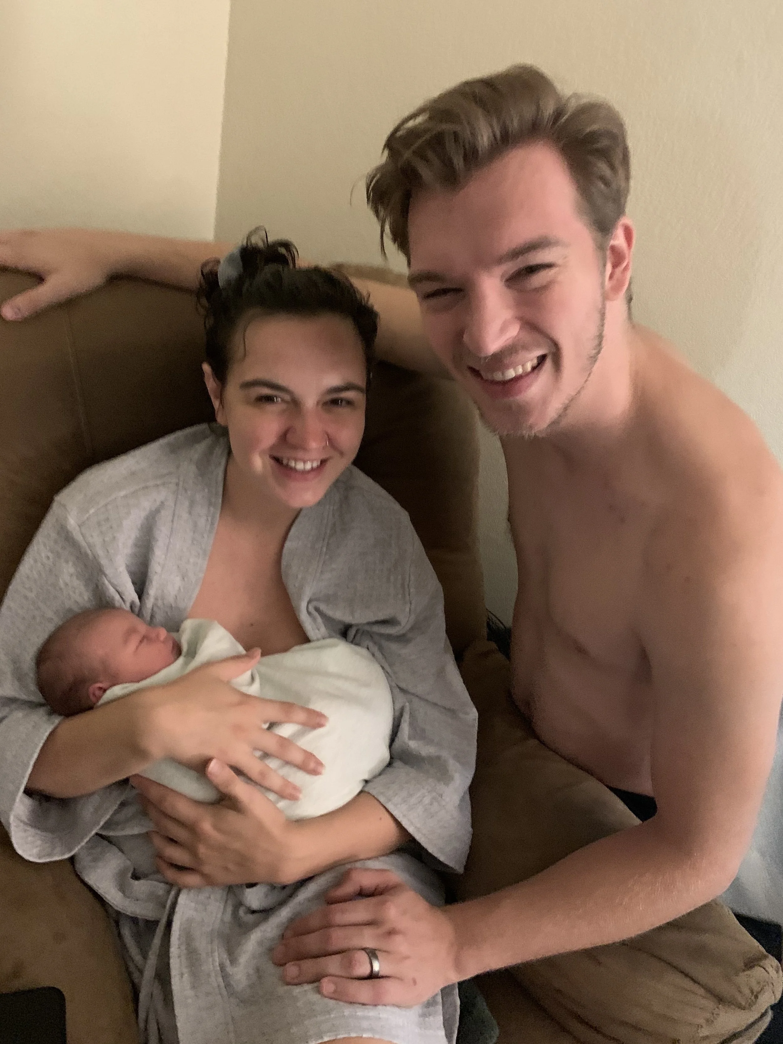 New mother in hospital gown holding a newborn baby wrapped in a blanket, smiling; father shirtless, leaning in with a happy expression, in a cozy indoor setting.