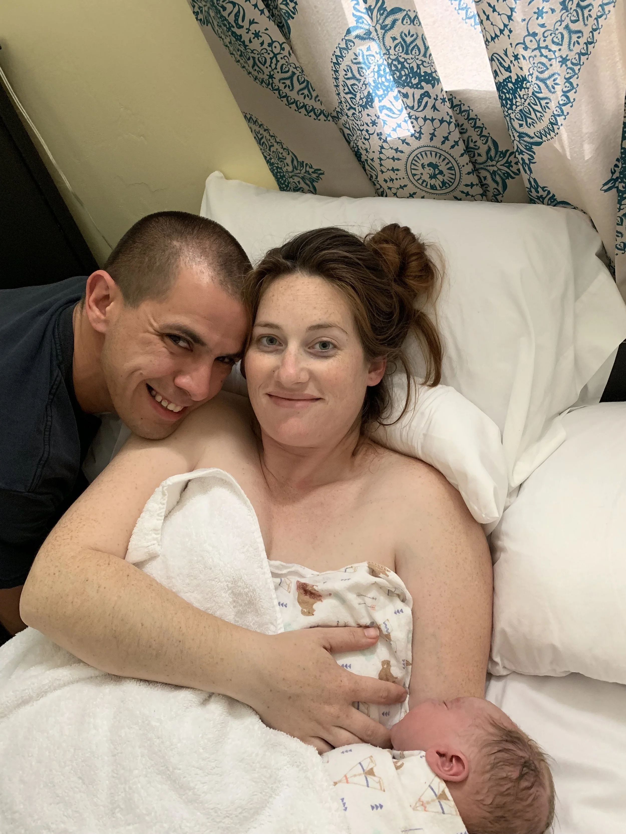 A woman holding a newborn baby in bed, with a man hugging her and smiling. The woman has red hair and freckles, the man has short brown hair, and the baby has dark hair and is wrapped in a blanket.