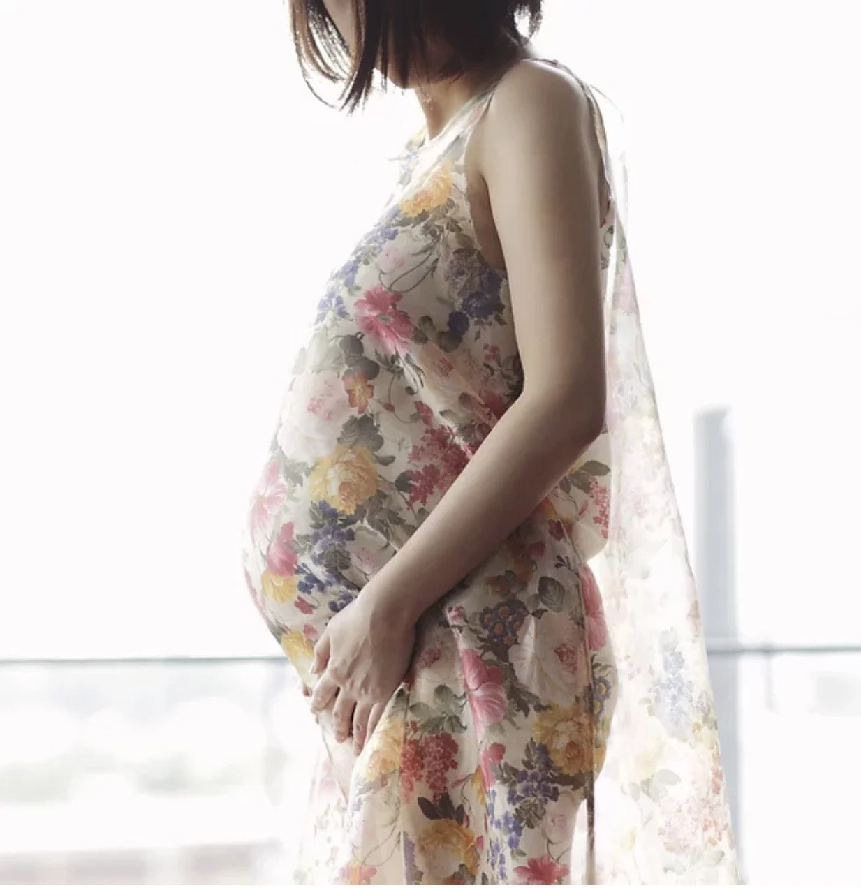 Pregnant woman wearing a floral dress standing near a window.