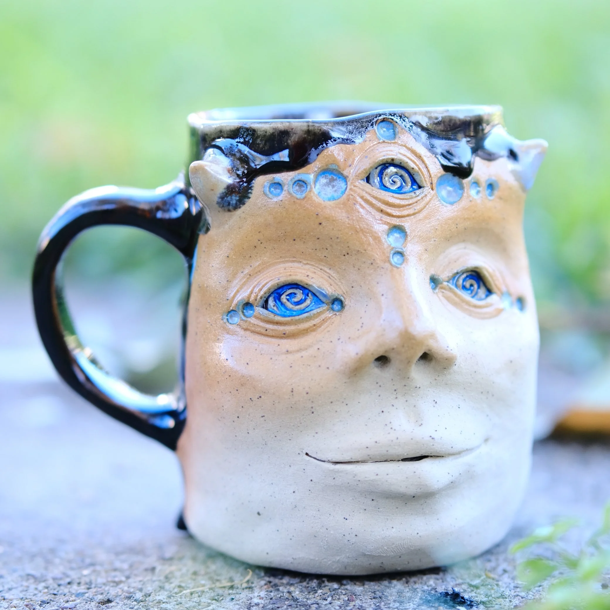 Hand-sculpted black ceramic shadow fairy mug with protruding horns, three blue spiral eyes, and drips of amber glaze over a dark textured surface.