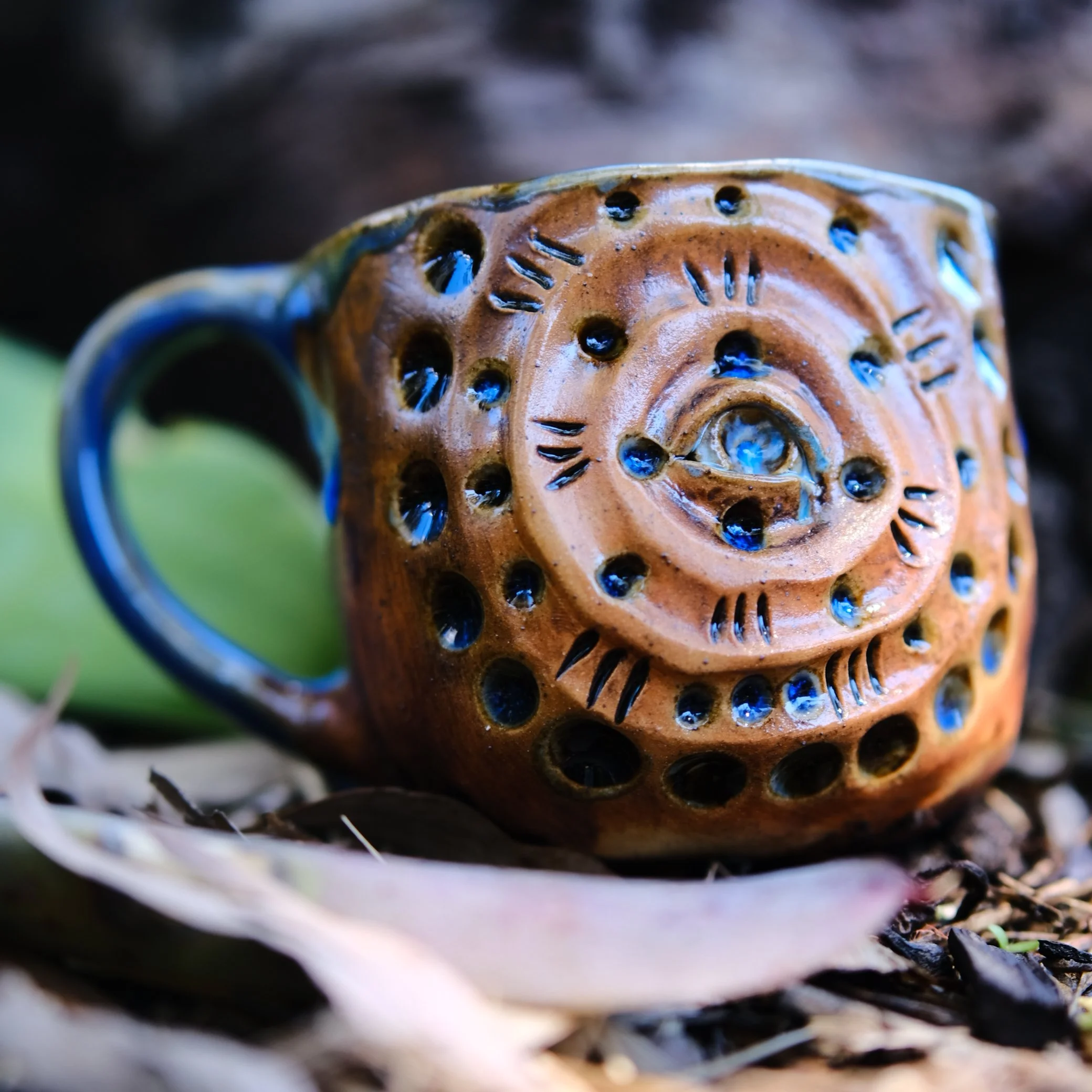 Hand-built tribal ceramic mug with a central carved sacred eye motif and earthy brown textures, photographed in a natural woodland setting.