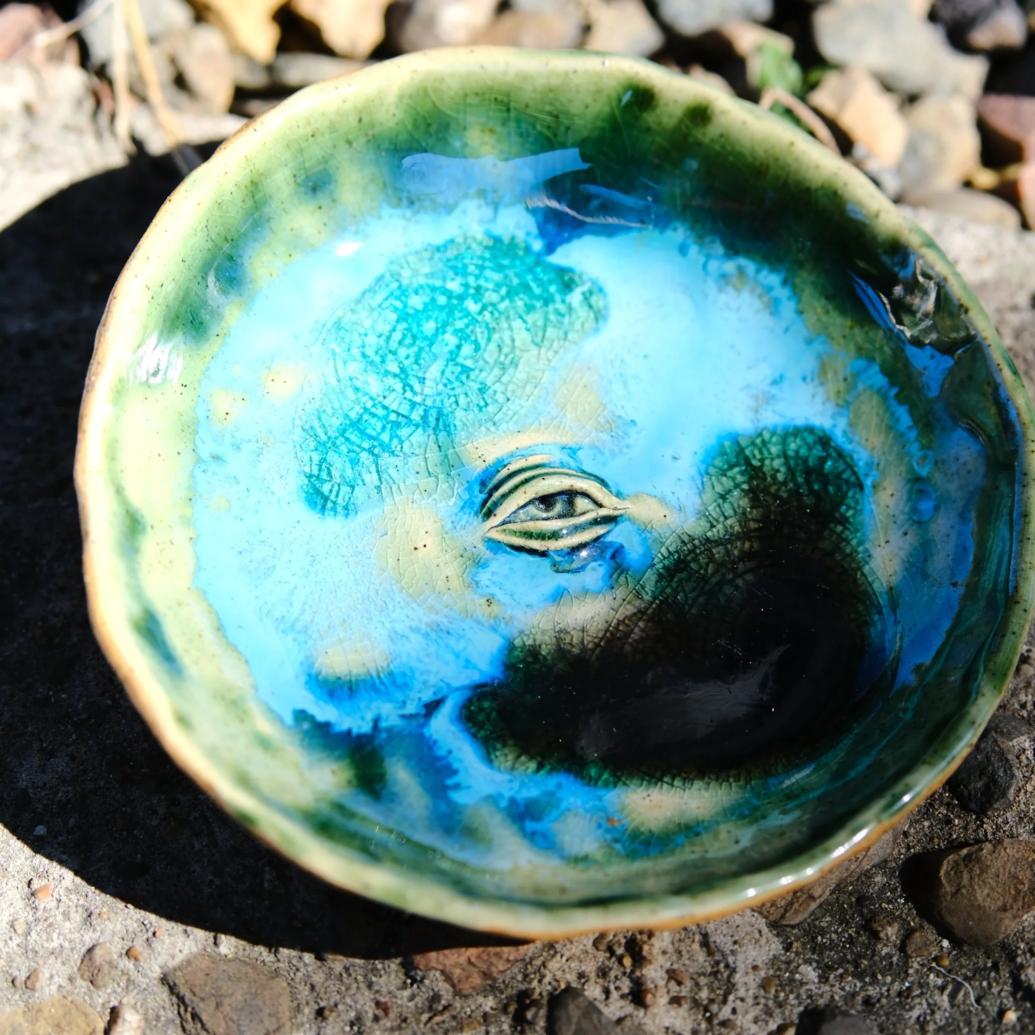 lose-up of a circular ceramic trinket dish with a central eye motif and a distinct blue and green crackle-effect glaze resembling ocean water.