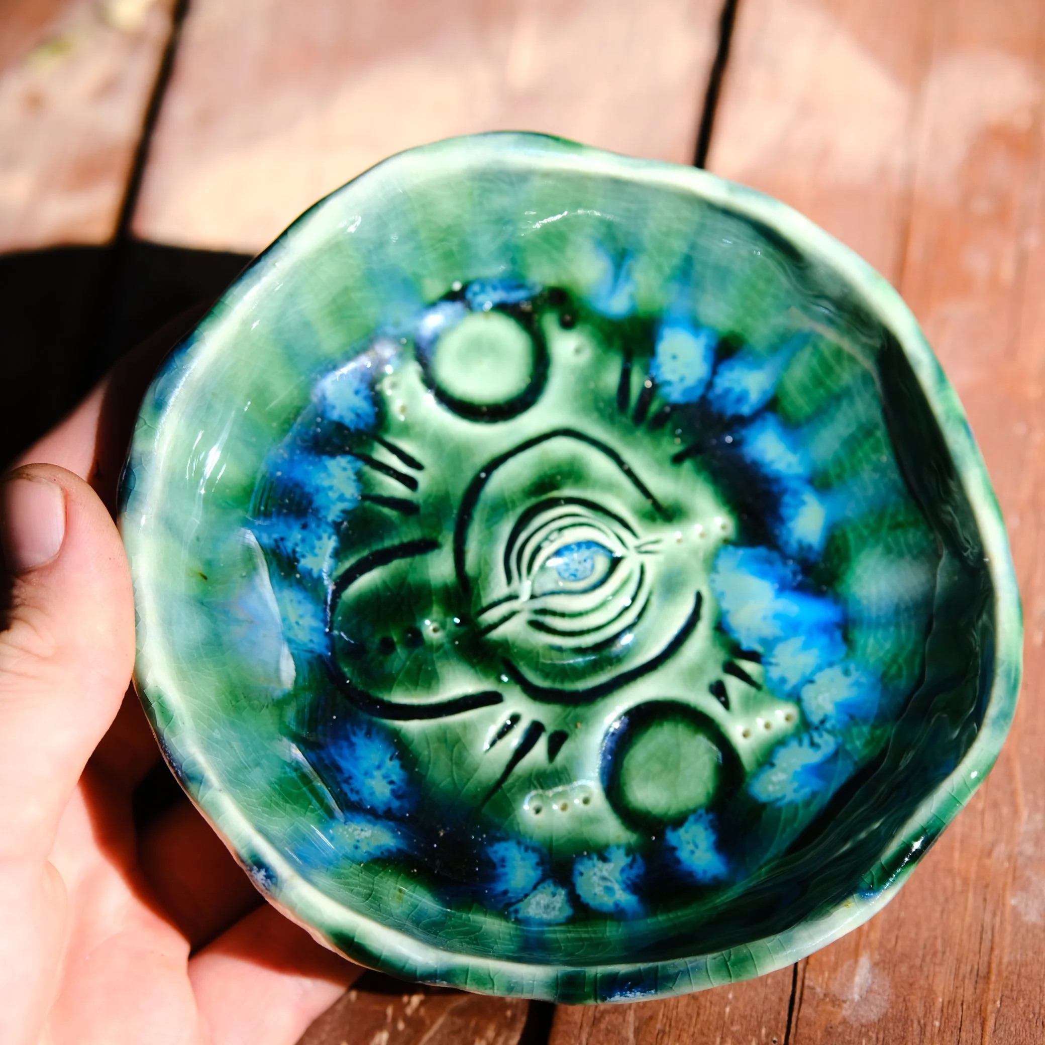 Handmade circular ceramic trinket dish with a central all-seeing eye motif surrounded by tribal patterns in vibrant blue and forest green glazes.
